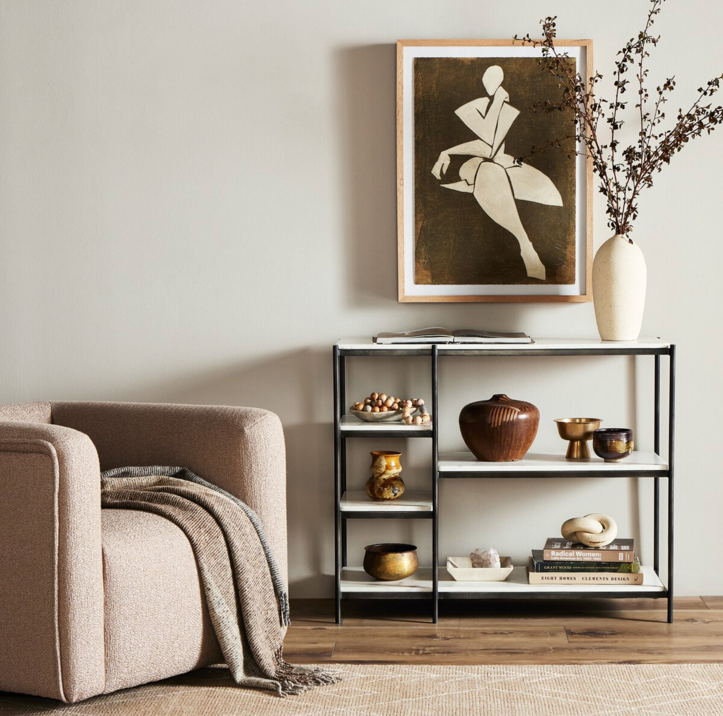 A cozy living room corner with a beige armchair, a blanket, and the Hammered Metal Console Table styled with books, decorative objects, a tall vase with branches, and abstract art displayed on the wall.
