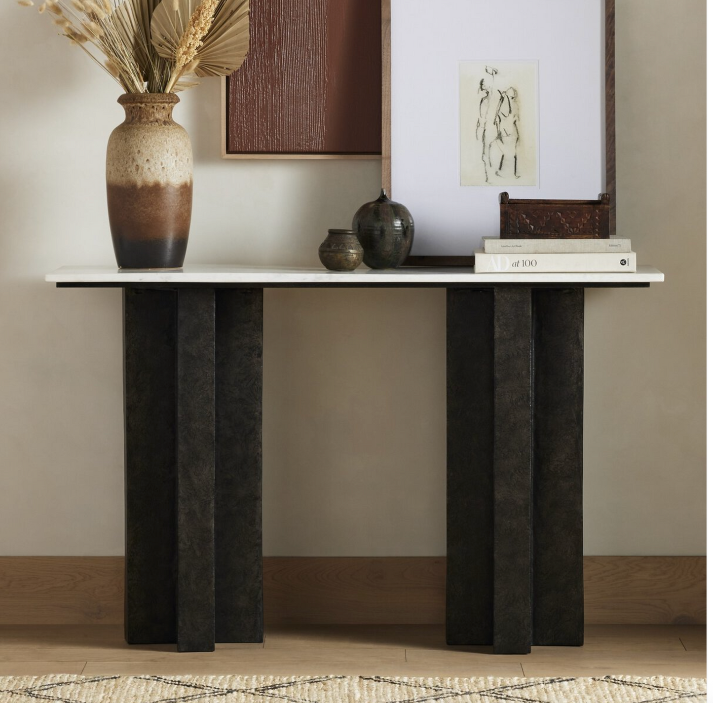 The Marble Raw Black Console Table, featuring a rectangular top and four dark cast aluminum legs, displays ceramic vases, dried plants, stacked books, and framed art against a light wall and wooden floor.