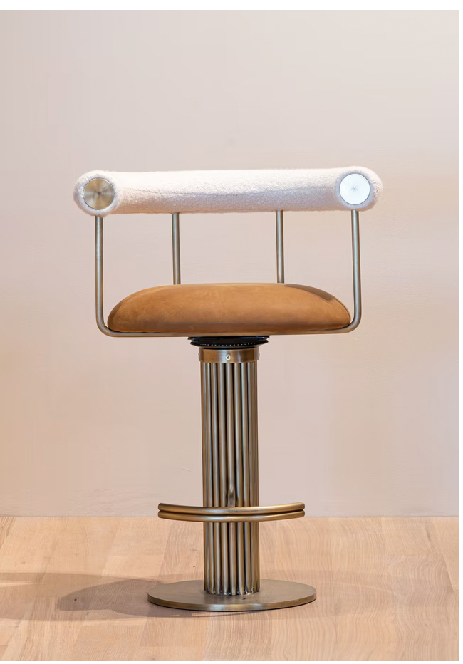The Brass Swivel Bar Stool features a tan cushioned seat, metal frame, white cylindrical backrest, and ribbed metallic pedestal base, shown on a light wooden floor against a plain wall.
