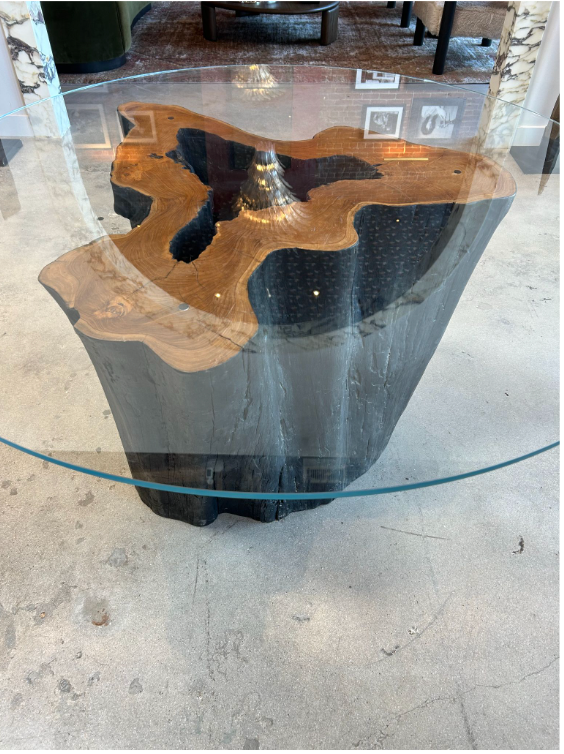 The Teak Trunk Table With Glass Top features a round Starphire glass surface atop an old growth teak trunk base with textured grain and dark crevices, perfect for modern rooms with concrete floors.