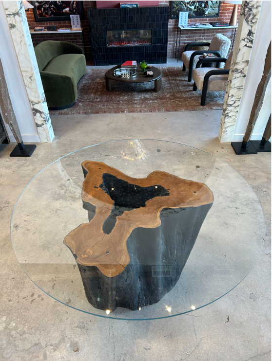 The Teak Trunk Table With Glass Top, featuring a round Starphire glass top and base crafted from old growth teak, stands on a concrete floor in a modern living room with green patterned chairs and a black fireplace in the background.