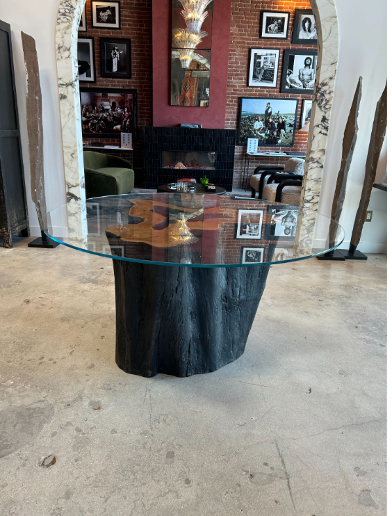 The Teak Trunk Table With Glass Top, featuring a round glass surface on a black tree trunk base, stands in a modern room with brick walls, framed photos, a marble archway, fireplace, green sofa, and stylish decor in the background.