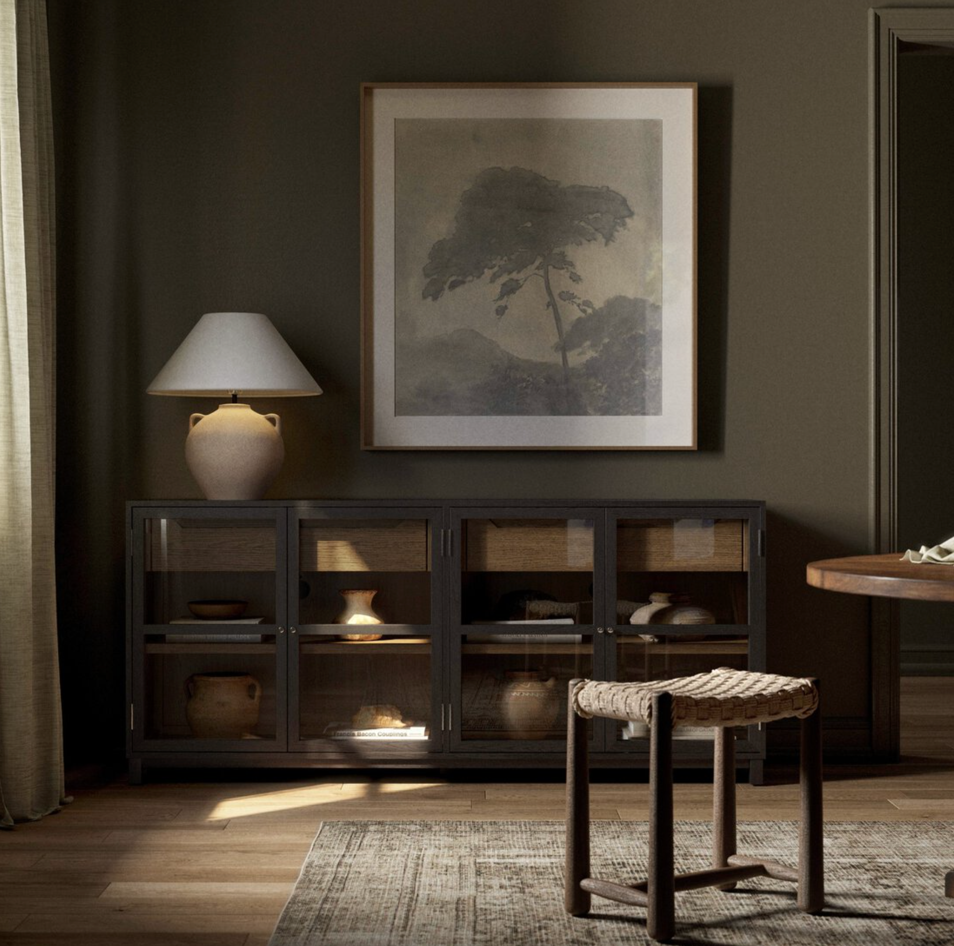A cozy, neutral-toned room with sunlight streaming in, featuring a wicker stool, an Oak Frame Large Sideboard with pottery inside, a table lamp, and framed tree artwork on the wall above the sideboard.