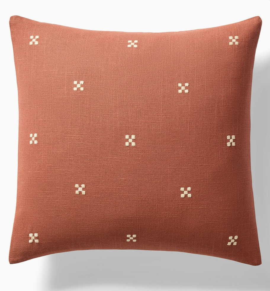 The Hand Embroidered Terracotta Pillow is a 20" square pillow in rust color, made of textured fabric and decorated with small, evenly spaced off-white embroidered squares for a simple, elegant look.