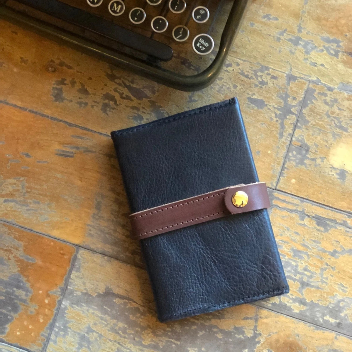 A Writer's Journal with a black leather cover, brown strap, and gold button rests on a worn wooden surface near a vintage typewriter—creating the perfect scene for an elegant corporate gifting moment or as a custom branded keepsake.