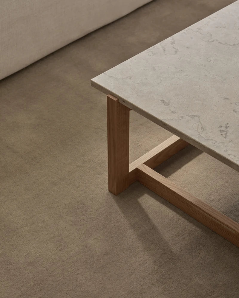 A close-up of a modern coffee table with a light stone top and wooden legs on the Sand Wool Rug, with a beige sofa in the background.