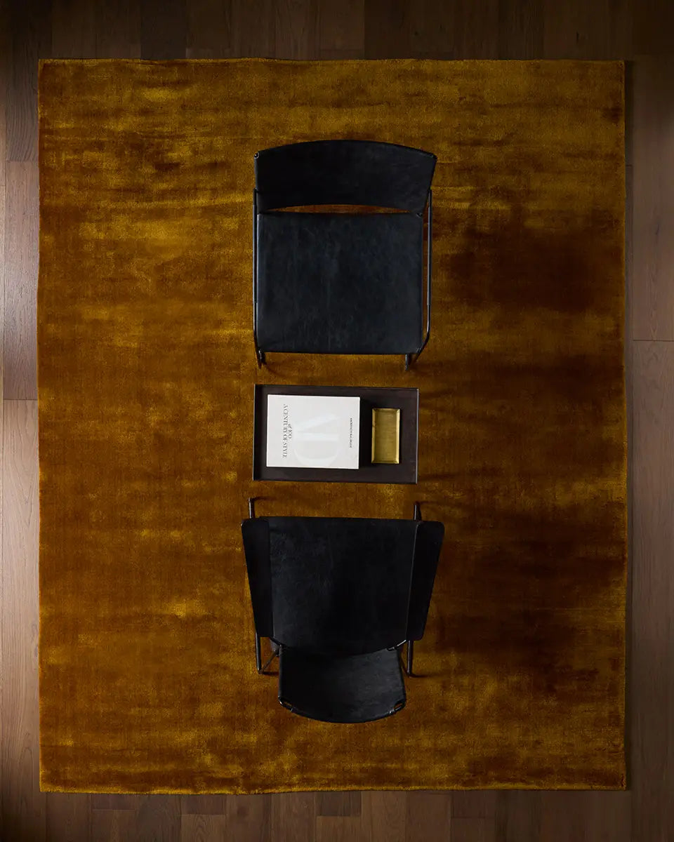 Overhead view of two black chairs facing each other across a small table with a book and a square tray, all arranged on the elegant Handwoven Golden Wool Rug, crafted from premium New Zealand wool and perfectly sized at 8x10 feet, atop a wooden floor.