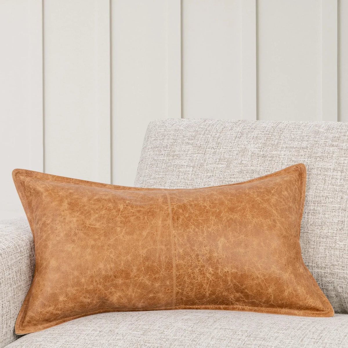 A Leather Lumbar Pillow in brown, measuring 14″ x 26″, rests on a light gray textured sofa, with vertical beige wall panels in the background.