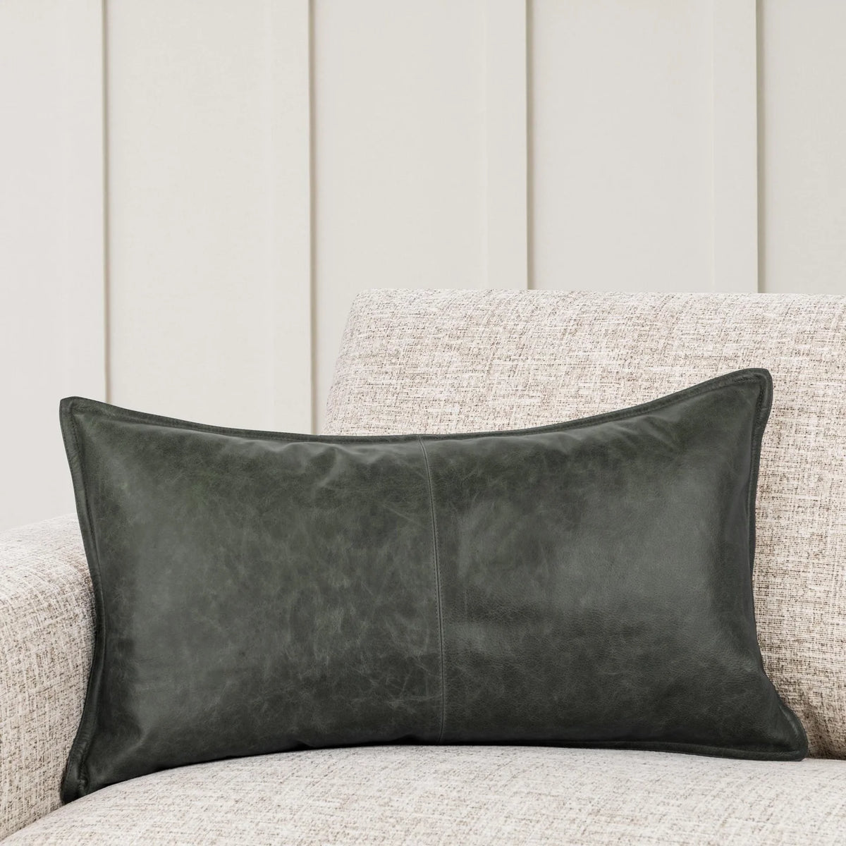 The Leather Lumbar Pillow, featuring a rectangular shape in dark green leather and a 14x26 pillow insert, rests on a light beige textured sofa in front of a cream-colored paneled wall.