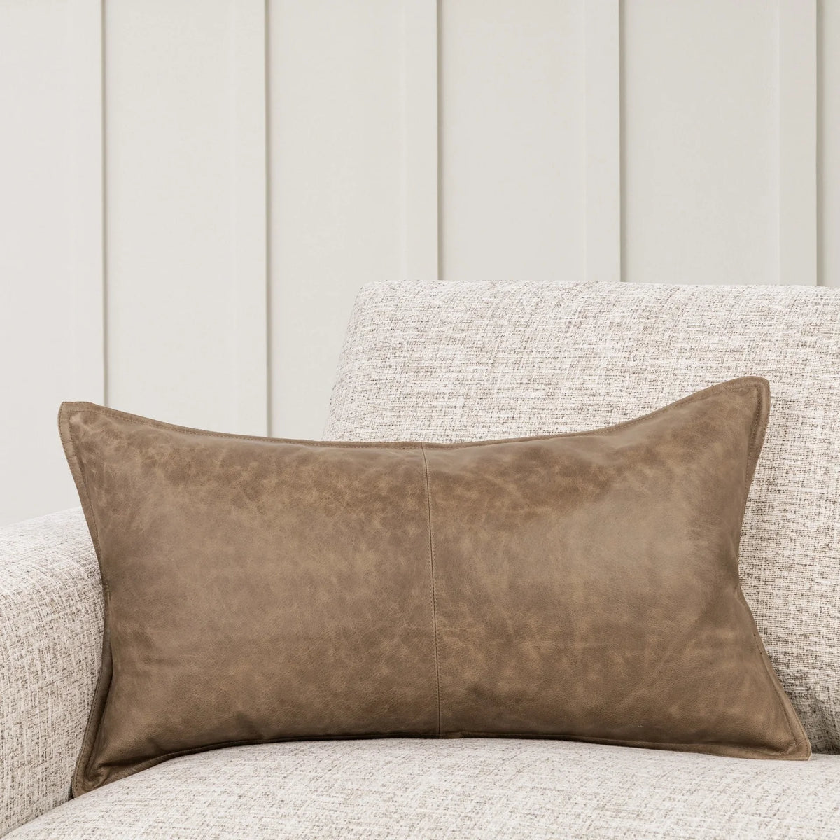 A Leather Lumbar Pillow in rectangular brown leather with a 14x26 pillow insert rests on the arm of a light-colored textured fabric sofa, with a cream paneled wall in the background.