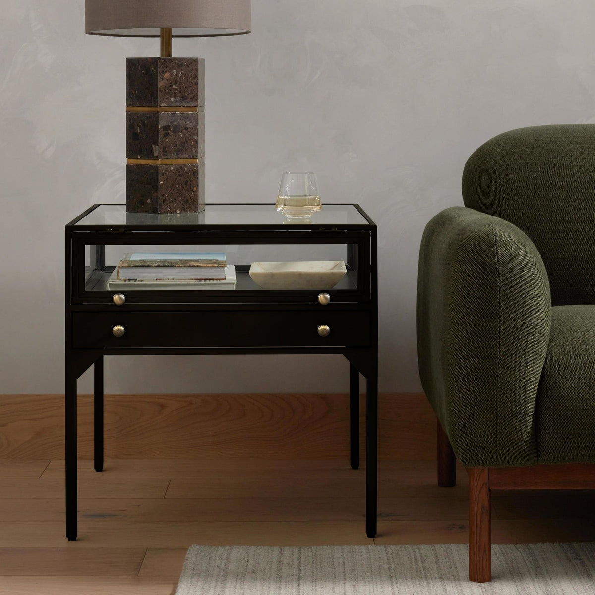 Black 24-inch Shadow box End Table featuring a glass-topped display compartment with two books and a small dish, a lower drawer, and a top surface holding a table lamp and a glass of water.
