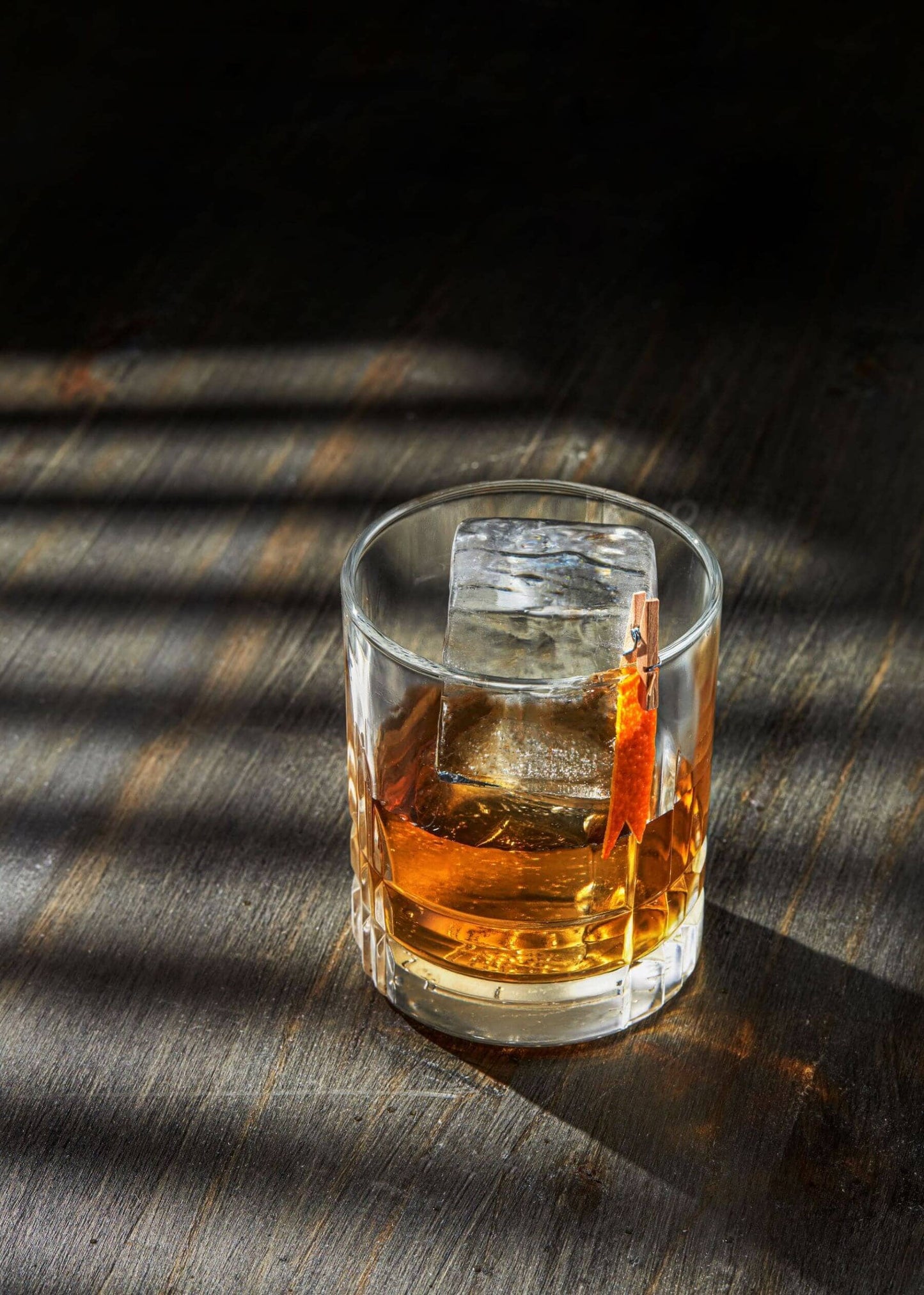 A glass of amber whiskey with a large, crystal clear ice cube—made using the Ice Cube Maker Tray (Eight 2" Squares)—and an orange peel garnish sits on a dark wooden surface, sunlight casting striped shadows across the scene.