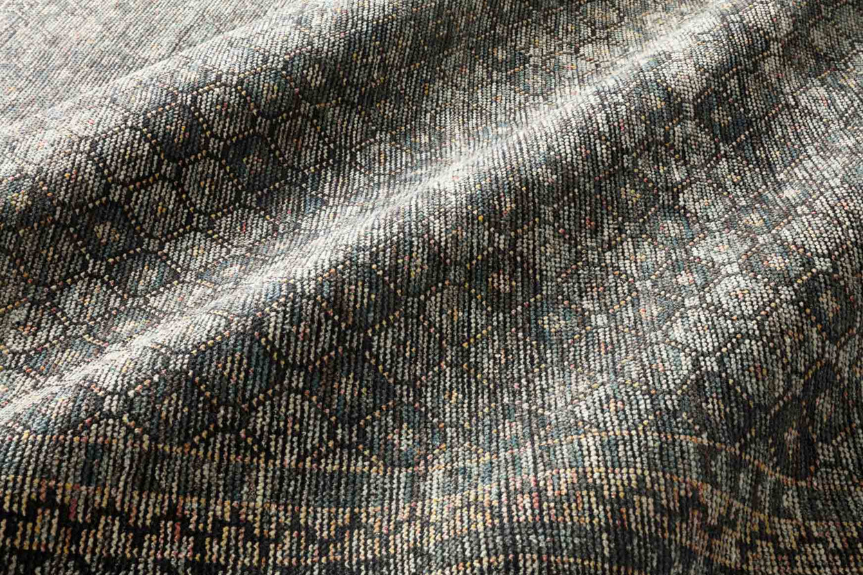 A close-up of the Ink|Turquoise Hand Knotted Rug 8'x10' reveals textured fabric with a subtle geometric pattern in shades of gray, black, and muted gold, gently draped to highlight folds and shadows.