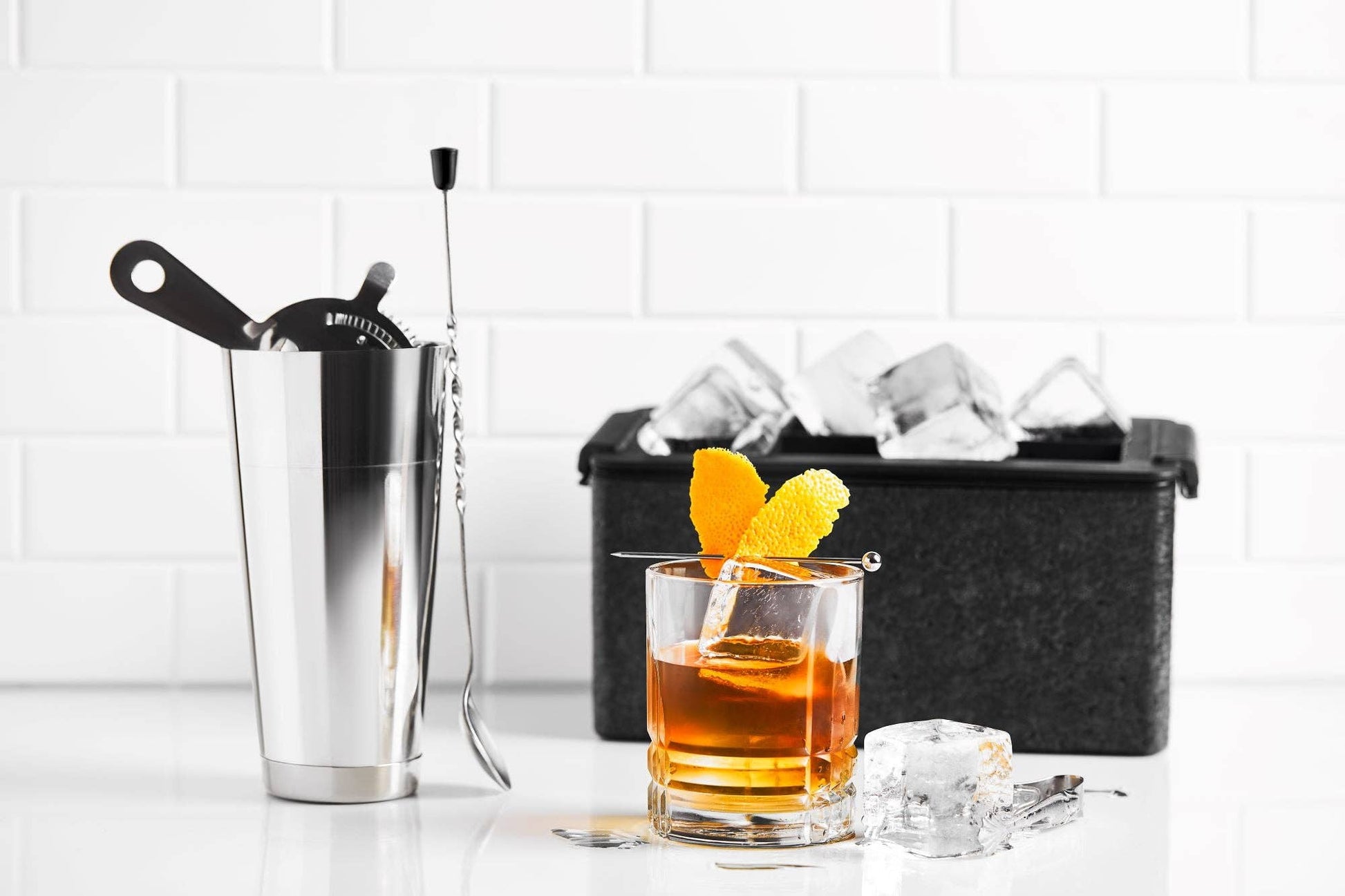 An Ice Cube Maker Tray - Eight 2" Squares Ice Cubes (1pc), cocktail shaker with bar tools, orange-garnished homemade cocktail in a glass, and black ice bin rest on a white surface against white tiled background.