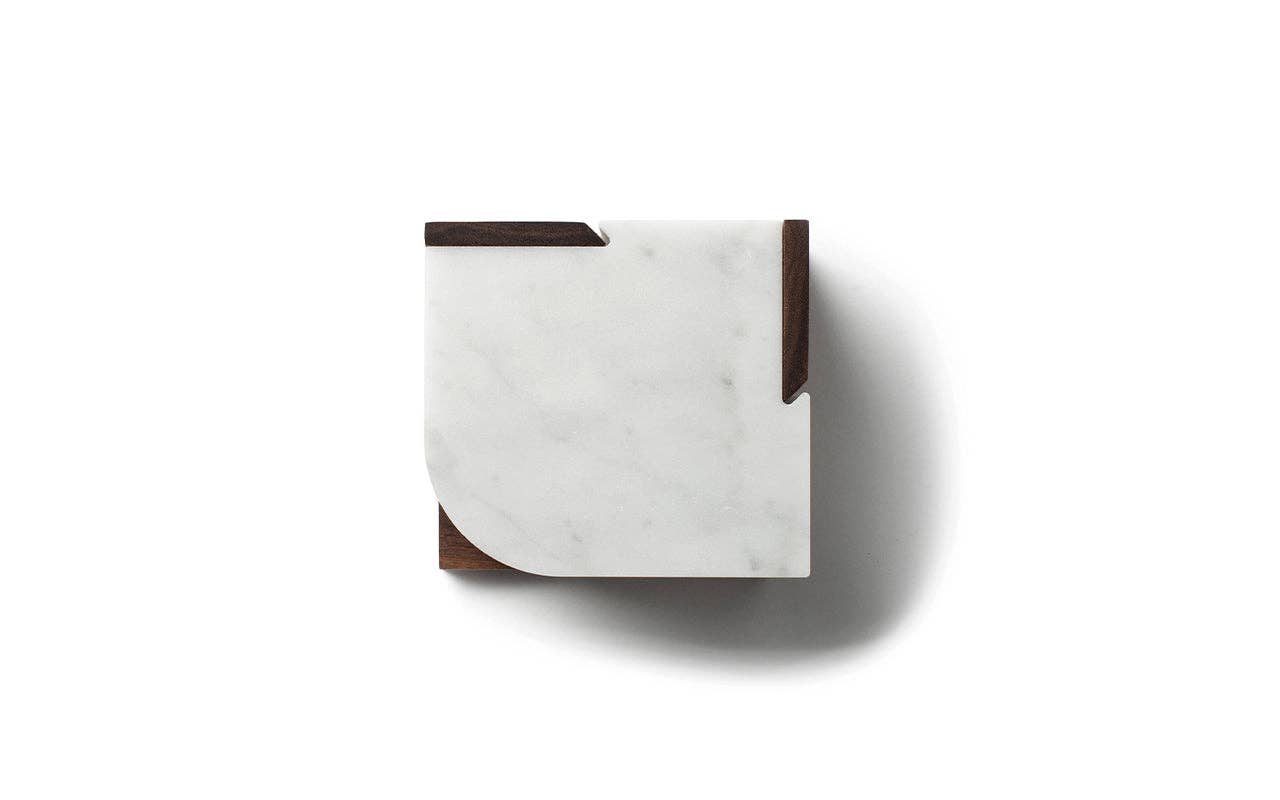 The Marble Coasters + Trivet features a square marble coaster with two wooden corner accents—one angular, one curved—casting a soft shadow on white, offering modern style perfect for coasters or as part of a trivet set.