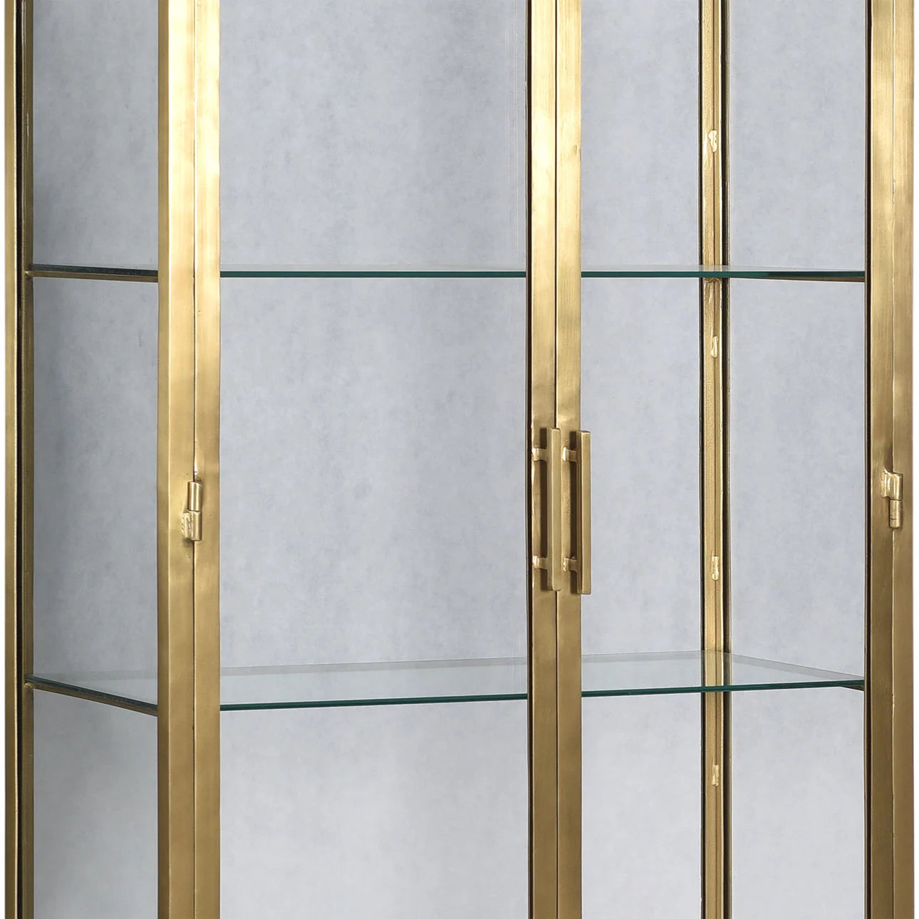 A close-up of the Brass Tall Display Cabinet featuring a gold metal frame, clear glass shelves, vertical handles on glass doors, and measuring 84" high by 32" wide. The background is plain light gray.