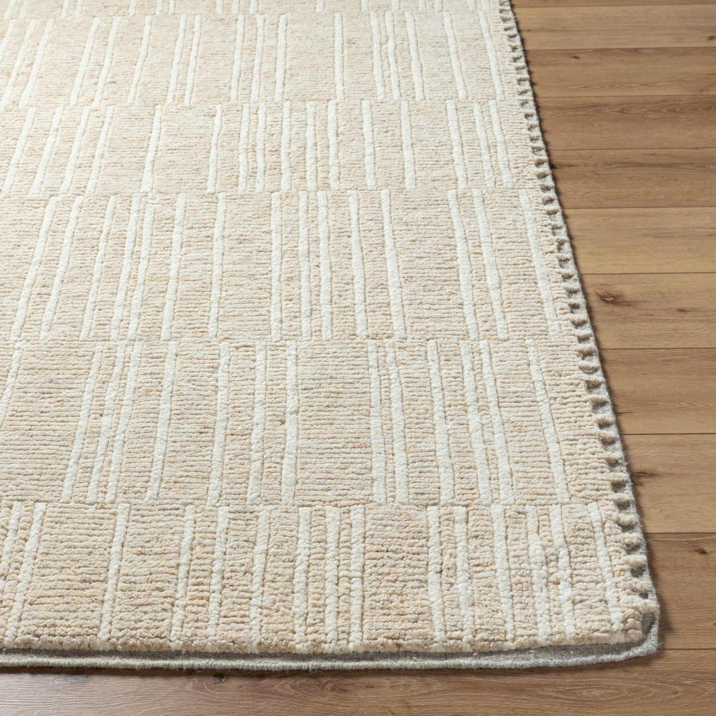 The Line Wool Rug- 8 x 10, made of beige textured 100% wool with a subtle geometric pattern, is shown on a wooden floor in a close-up of its corner and finely stitched edges.