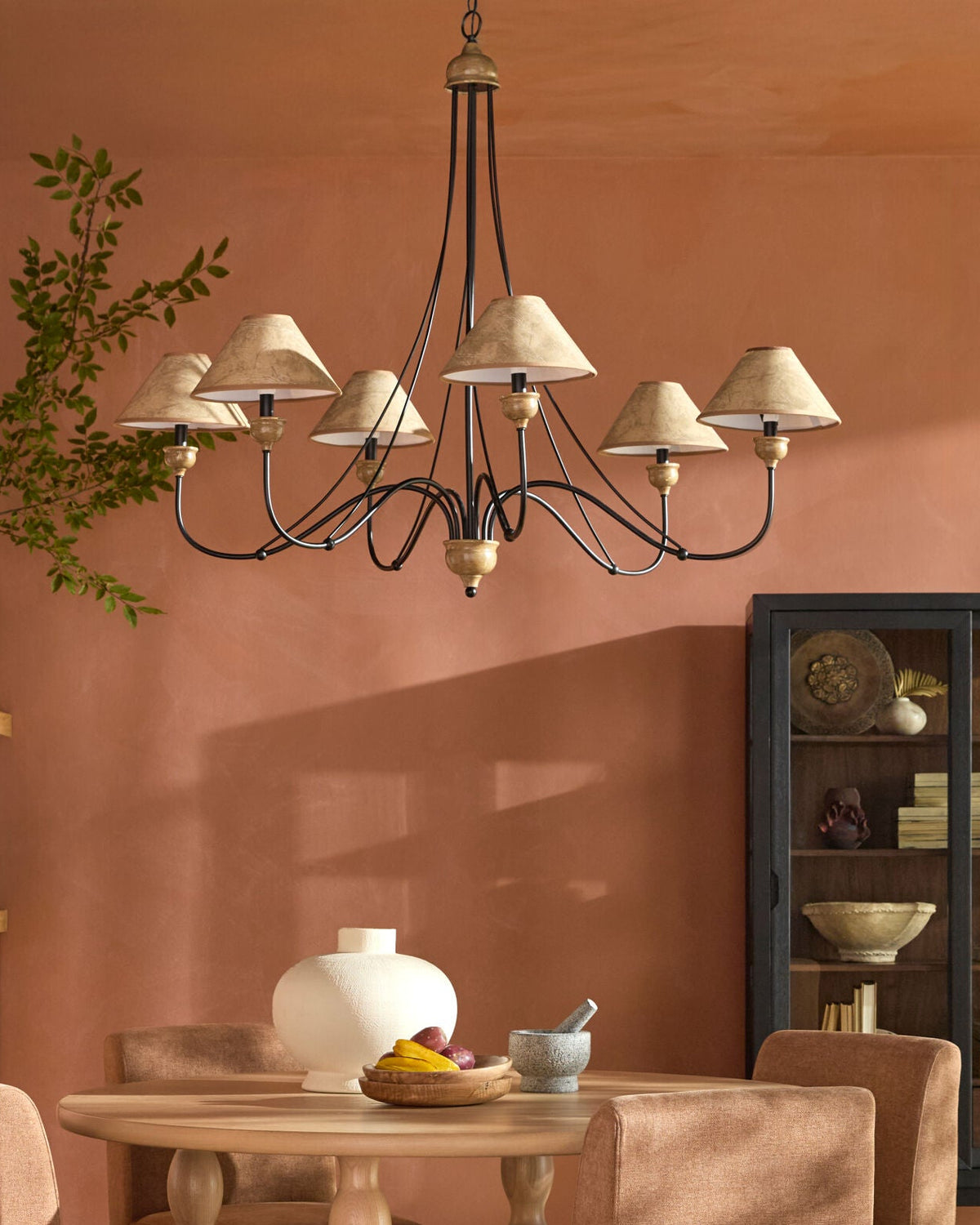An Iron and Leather Chandelier with six arms, black metal scrolls, and beige fabric shades hangs above a 41-inch square dining table.