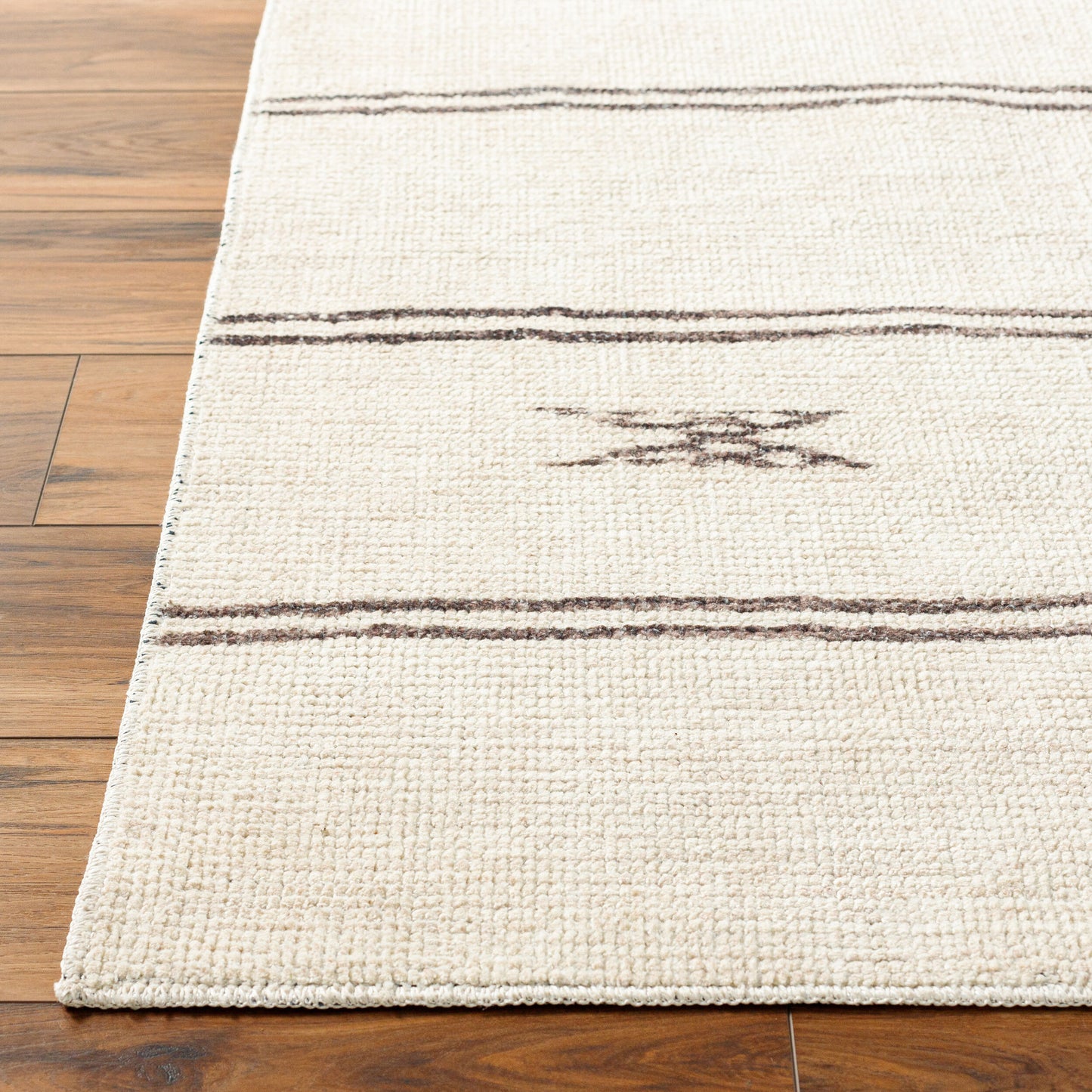 A close-up of the Turkish Tribal Rug - Ivory 7-10 x 10, a cream-colored machine woven rug featuring thin brown lines and a simple geometric brown pattern, placed on a wooden floor.