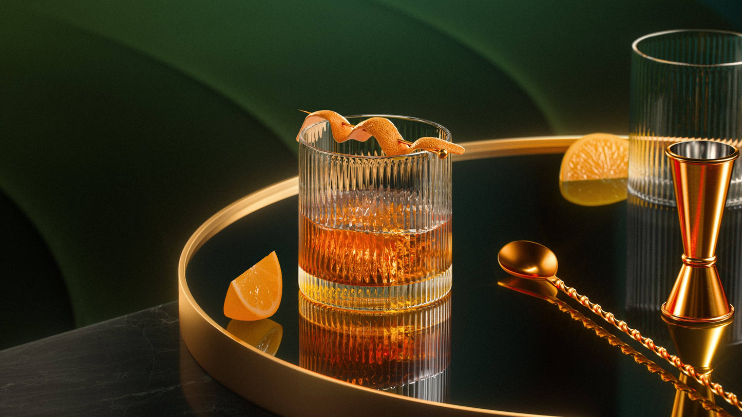 A glass of whiskey in a Vintage Straight Line Tumbler - 10 oz sits on a round black tray with gold edges, accompanied by a gold jigger, twisted bar spoon, and orange wedges against a green and gold backdrop.