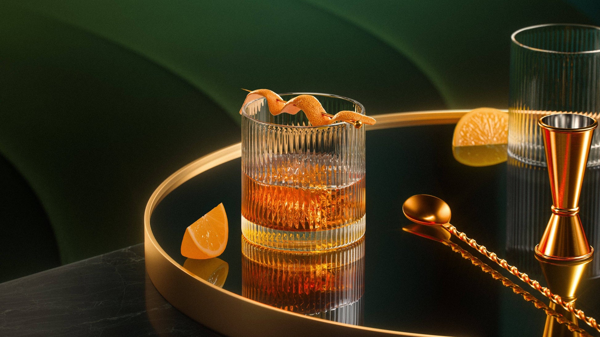 A glass of whiskey in a Vintage Straight Line Tumbler - 10 oz sits on a round black tray with gold edges, accompanied by a gold jigger, twisted bar spoon, and orange wedges against a green and gold backdrop.