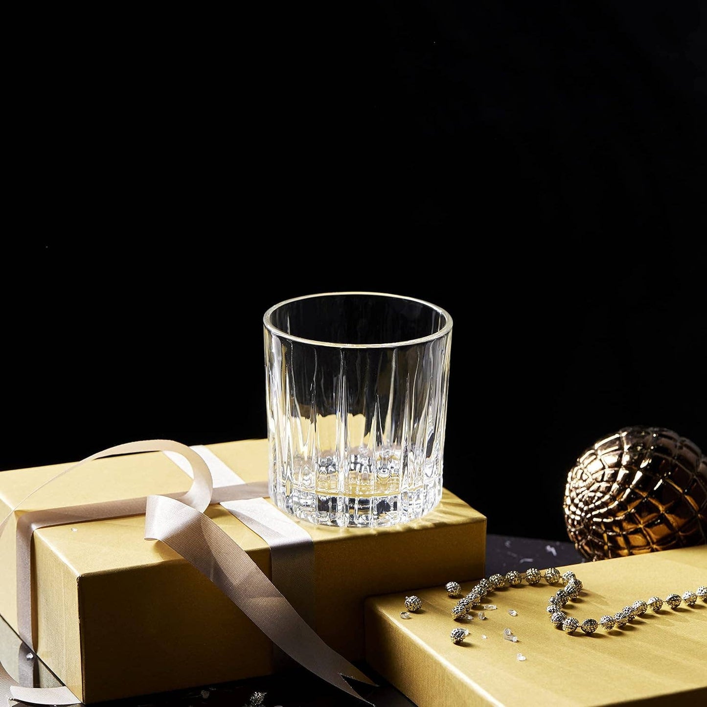A LEMONSODA Premium Double Rocks Whiskey Glass (12oz) rests atop a gold-wrapped gift box with a white ribbon, alongside a gold ornament and beaded necklace, against a dark backdrop—an ideal addition to any home bar.