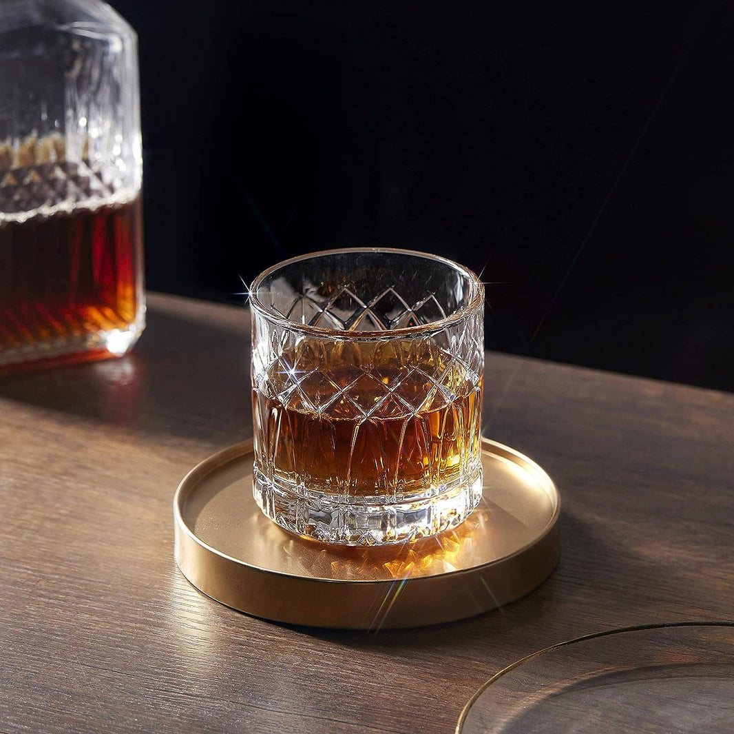 A LEMONSODA Premium Double Rocks Whiskey Glass (12oz) sits on a round gold tray atop a wooden surface, adding elegance to any home bar. A matching crystal decanter in the background enhances the allure of fine glassware.