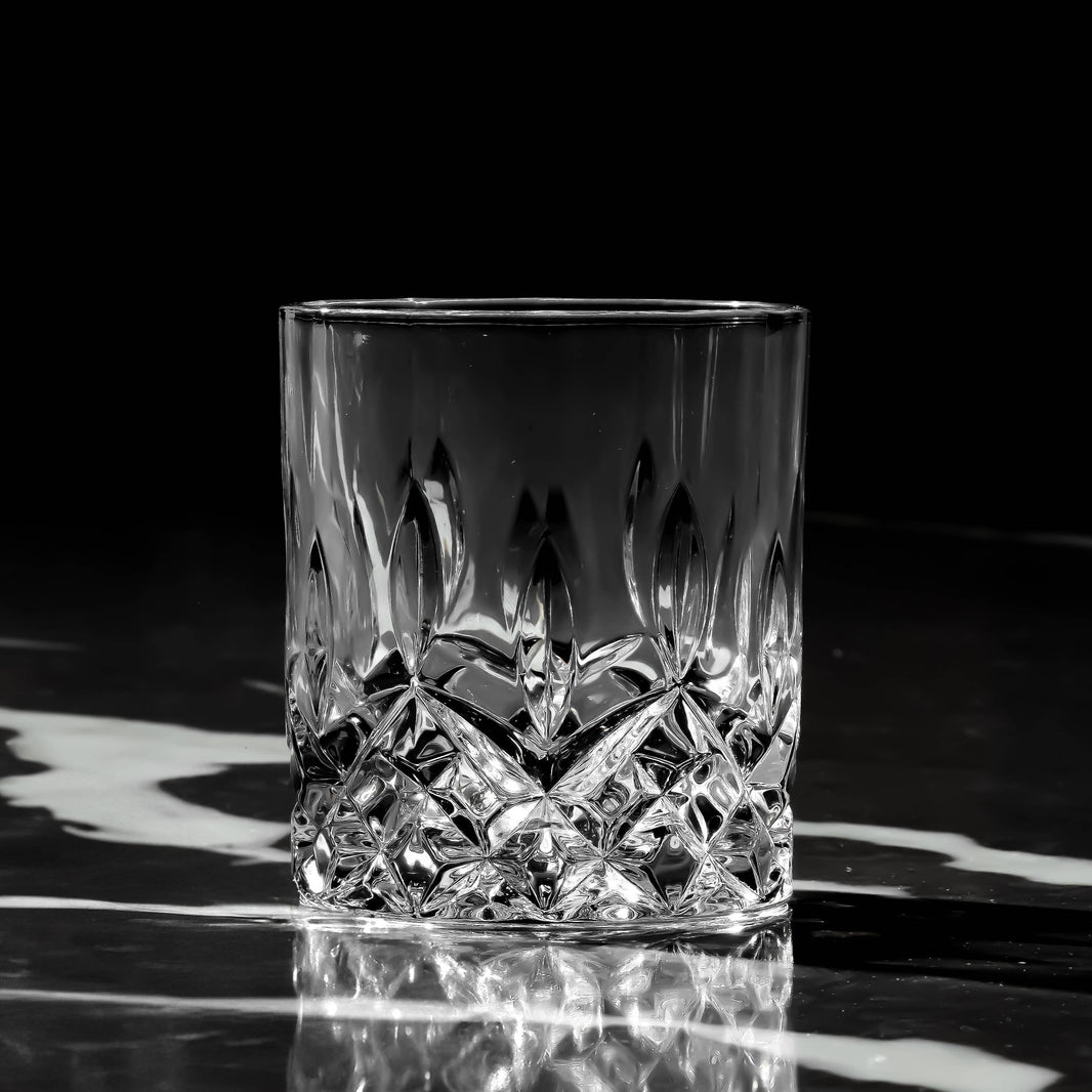 The Crystal Cut Old Fashioned Whiskey Glass 10oz (1pc) sits empty on a reflective marbled surface, its intricate patterns highlighted by a black background—an elegant, dishwasher-safe gift for any whiskey lover.