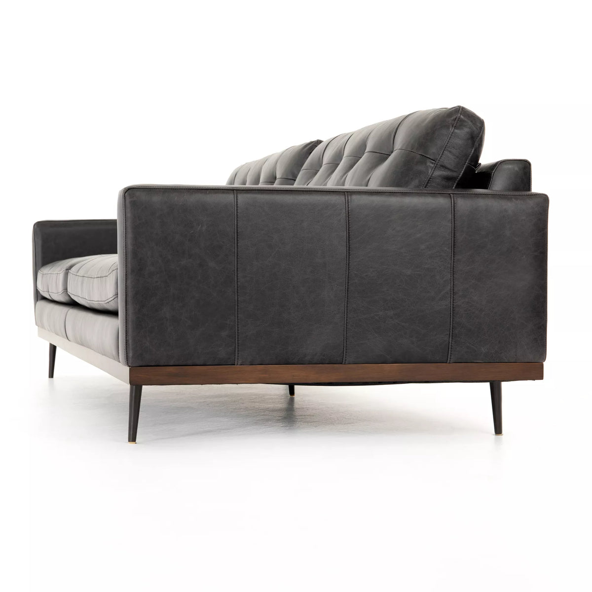 The Lexi Sofa is a modern 89-inch dark gray leather sofa featuring tufted back cushions, slim black metal legs, and a wooden base, shown from the side against a white background. Its elegant dimensions make the Lexi Sofa an ideal choice for contemporary living spaces seeking both style and comfort.