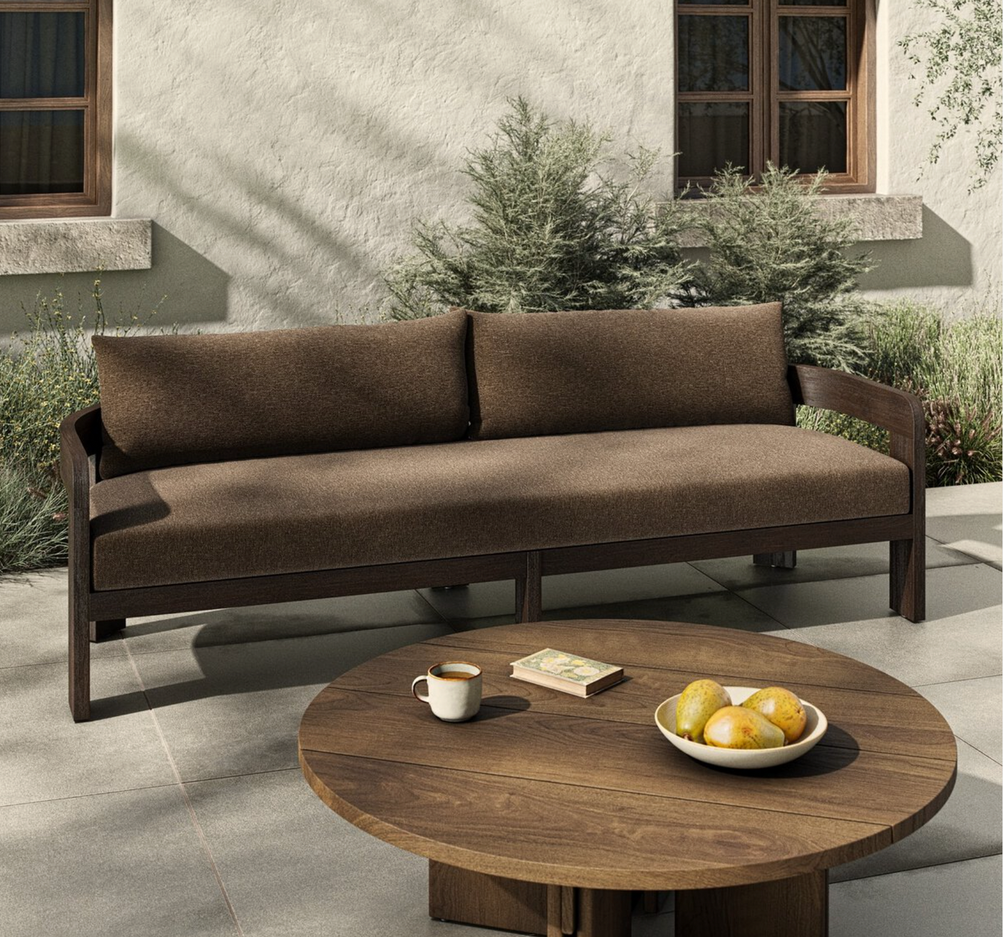 The Jackson Outdoor Sofa with teak framing is placed on a patio near green shrubs, paired with a round wooden table that holds lemons, a book, and coffee. Behind it is a beige wall with windows.