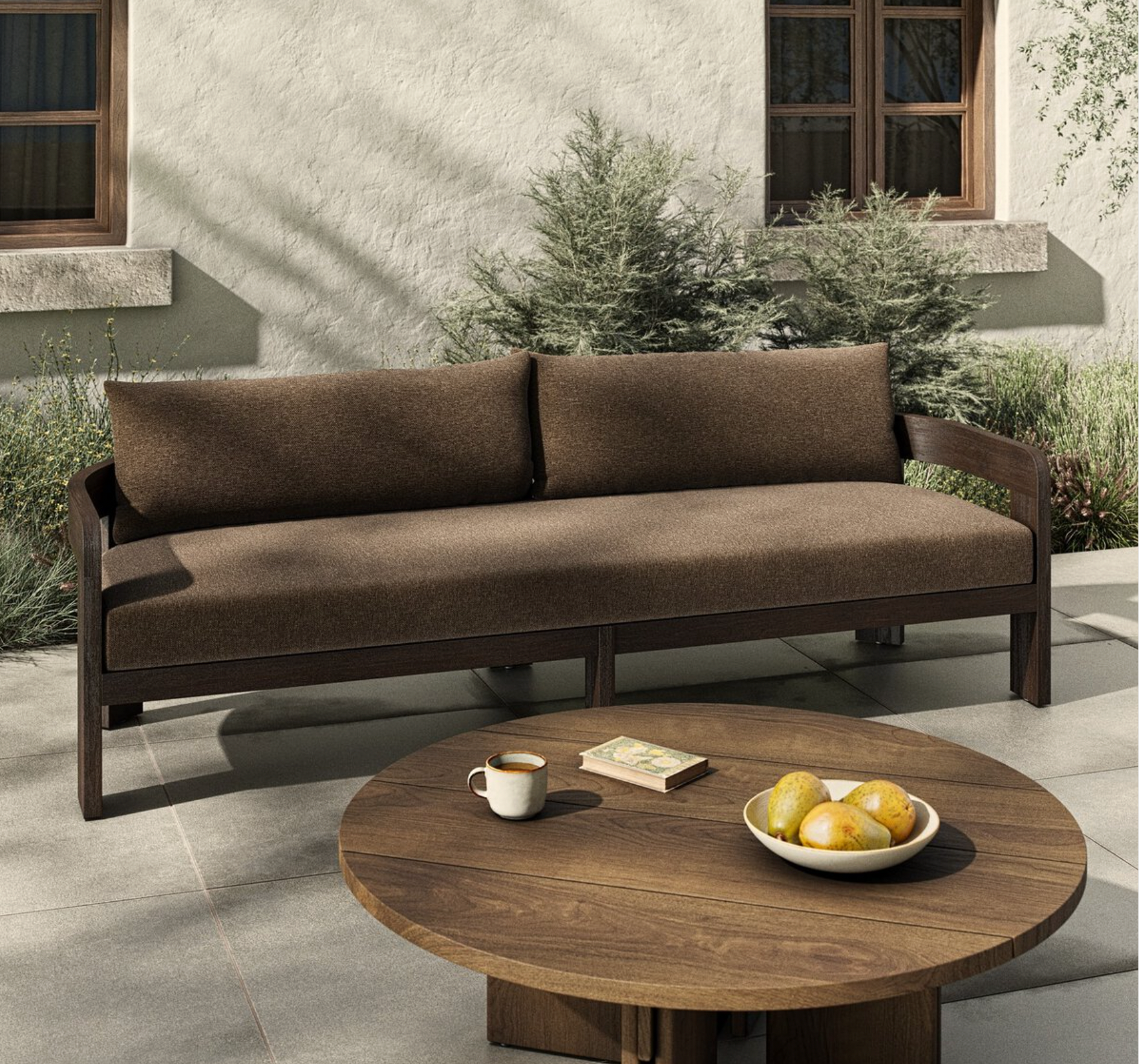 The Jackson Outdoor Sofa with teak framing is placed on a patio near green shrubs, paired with a round wooden table that holds lemons, a book, and coffee. Behind it is a beige wall with windows.