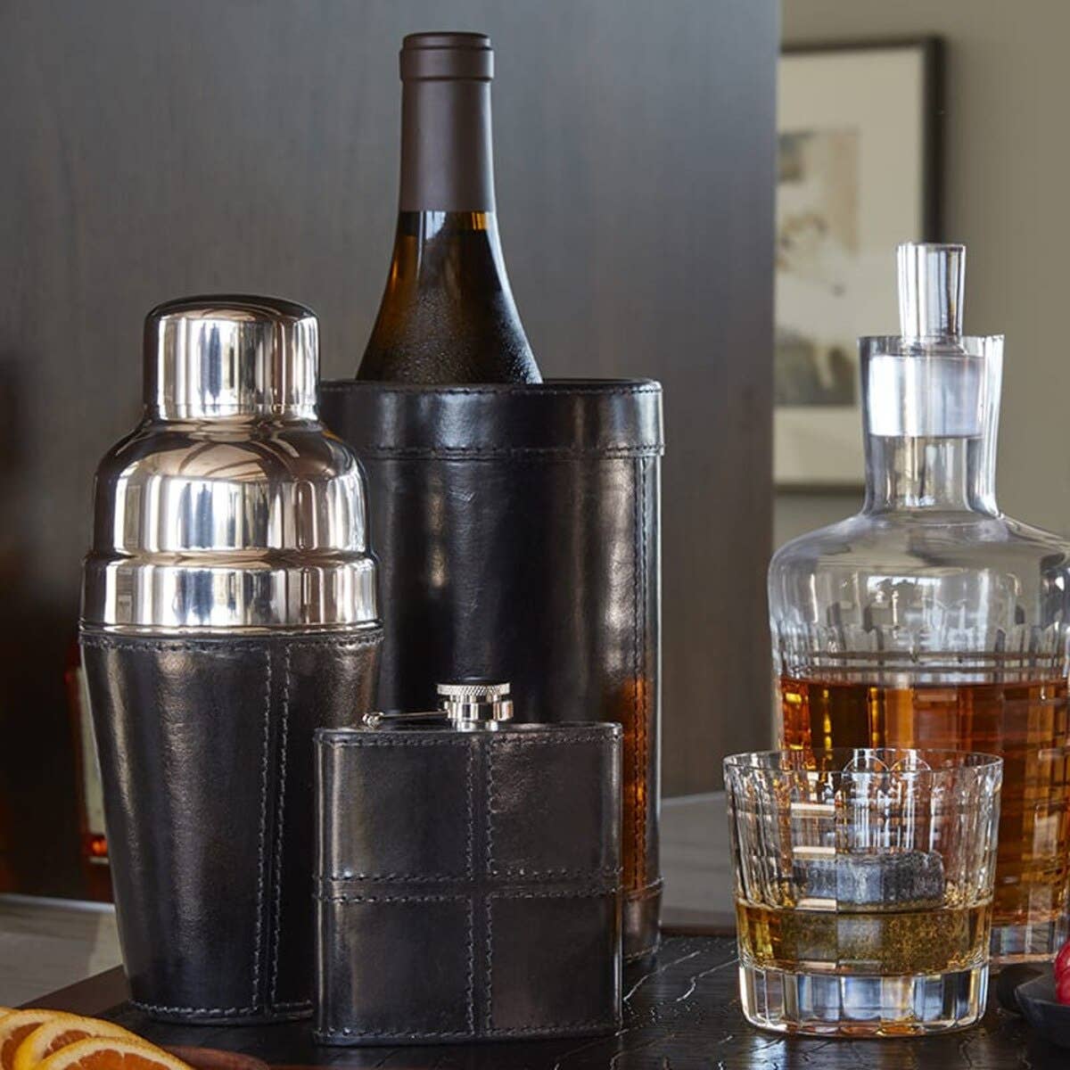 A stylish bar setup with the Leather Cocktail Shaker Black, a wine bottle in a leather holder, flask, whiskey decanter, and glass with ice—an elegant gift choice presented on a polished wooden surface.