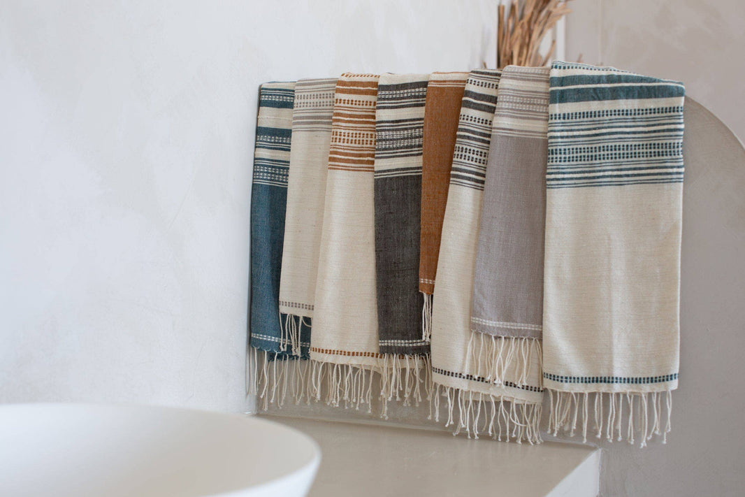 Five striped Handwoven Ethiopian Cotton Hand Towels in blue, brown, grey, and beige hang neatly on a bar by a white sink. Dried plants in the background highlight these beautifully crafted Fair Trade towels.