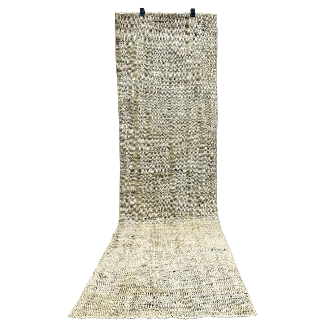 The Handmade Vintage Runner 3x9, a long beige Turkish piece with a subtle textured pattern, hangs vertically by two black clips at the top while its lower end rests spread on the floor.