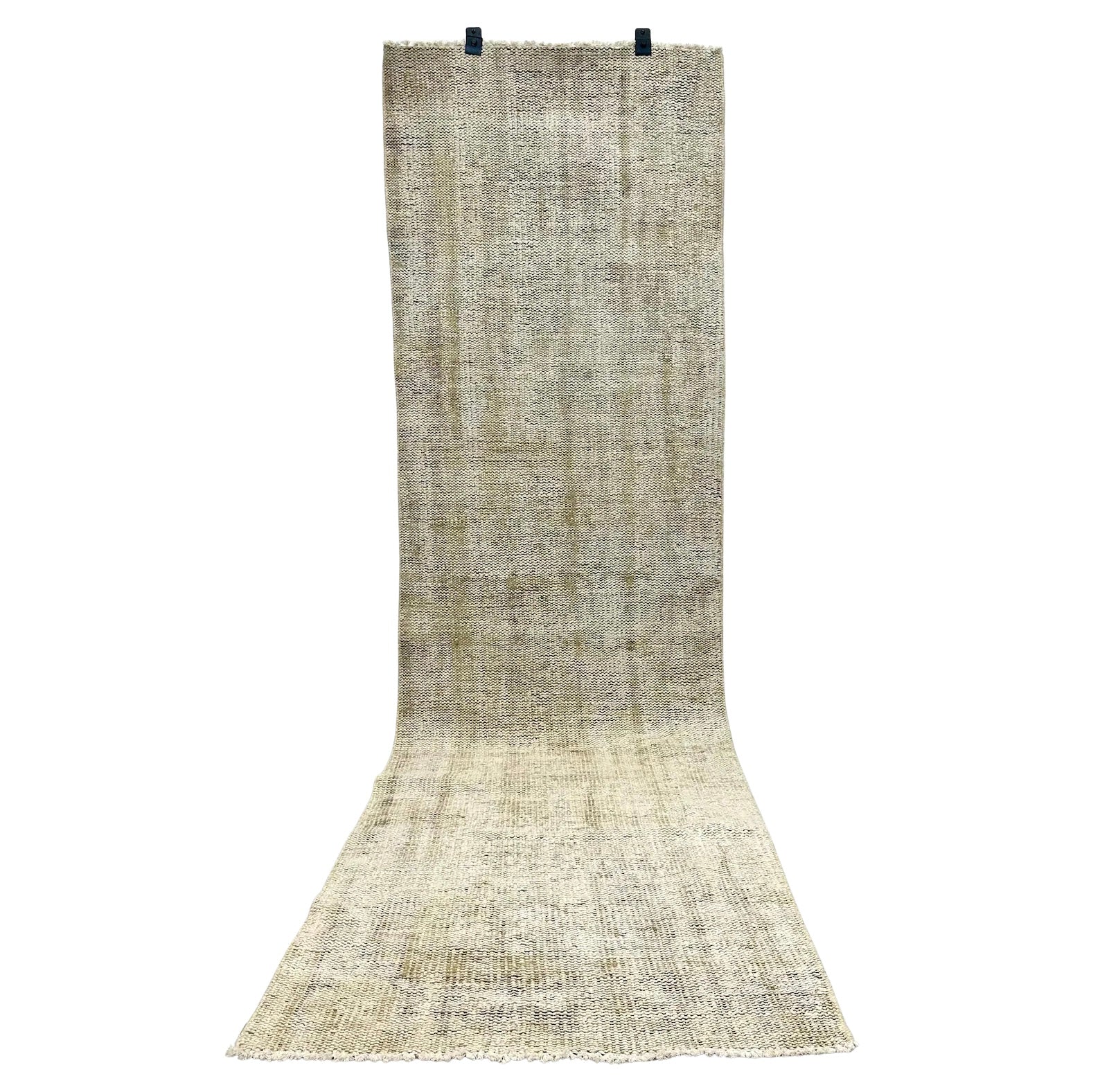 The Handmade Vintage Runner 3x9, a long beige Turkish piece with a subtle textured pattern, hangs vertically by two black clips at the top while its lower end rests spread on the floor.