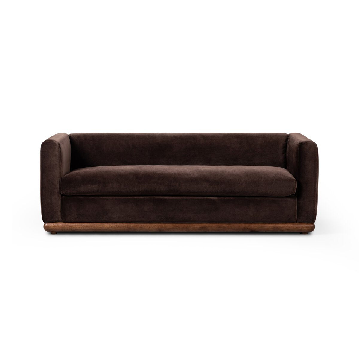 The Deco Sofa features a modern Art Deco design in dark brown velvet with a low profile, wide armrests, and a parawood plinth base, shown against a plain white background.