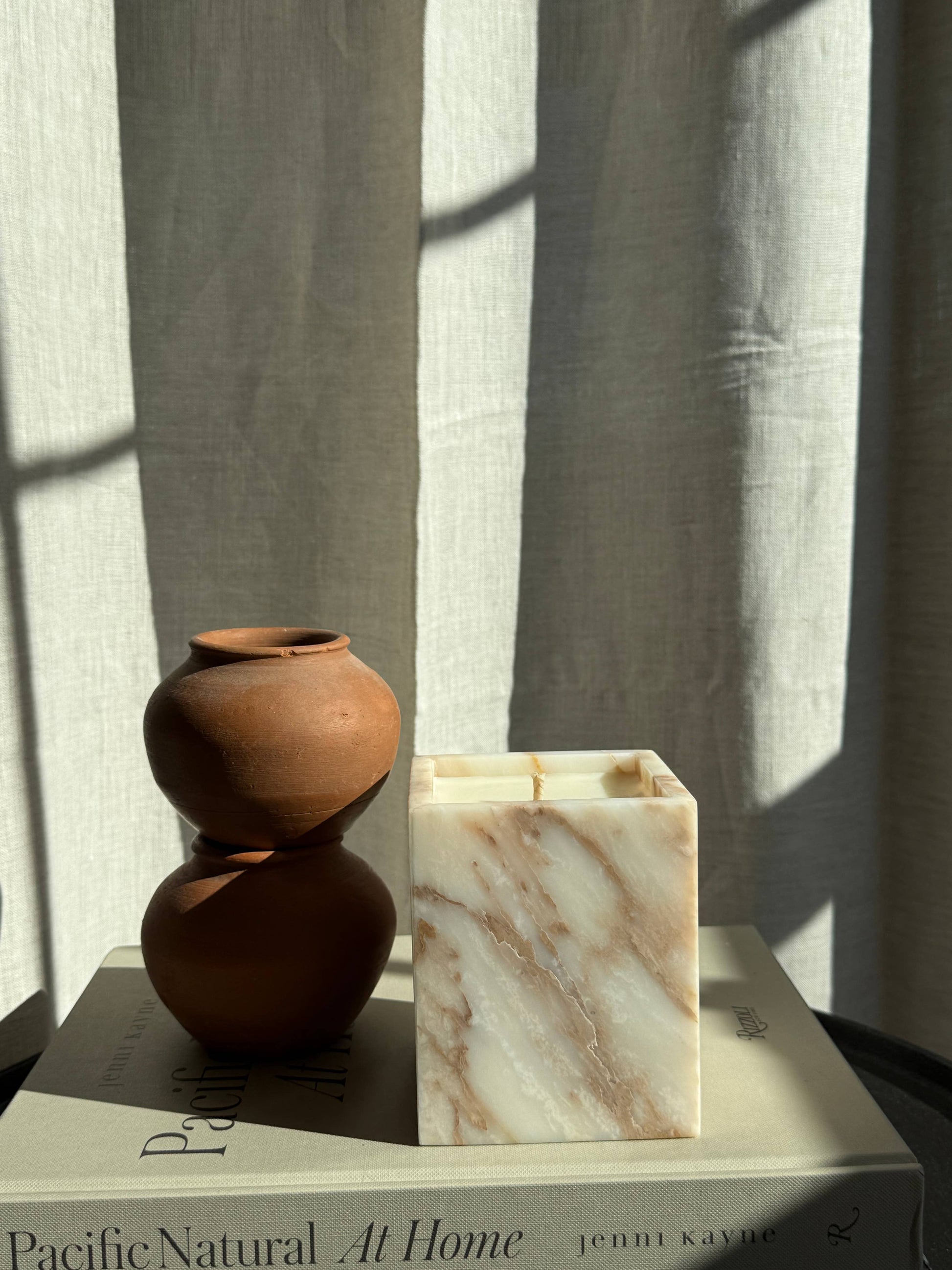 A small brown ceramic vase and a Marble Candles vessel rest on the book "Pacific Natural At Home" by Jenni Kayne, with sunlight and shadows falling across a neutral curtain in the background.