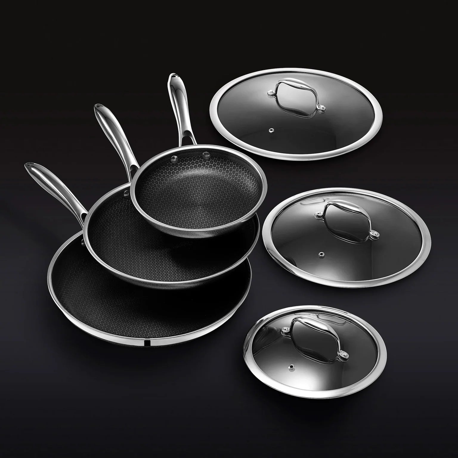 The Hexclad Fry Pan Set with Lids features three nonstick pans with metal handles, stacked together beside matching glass lids, all elegantly arranged against a black background.