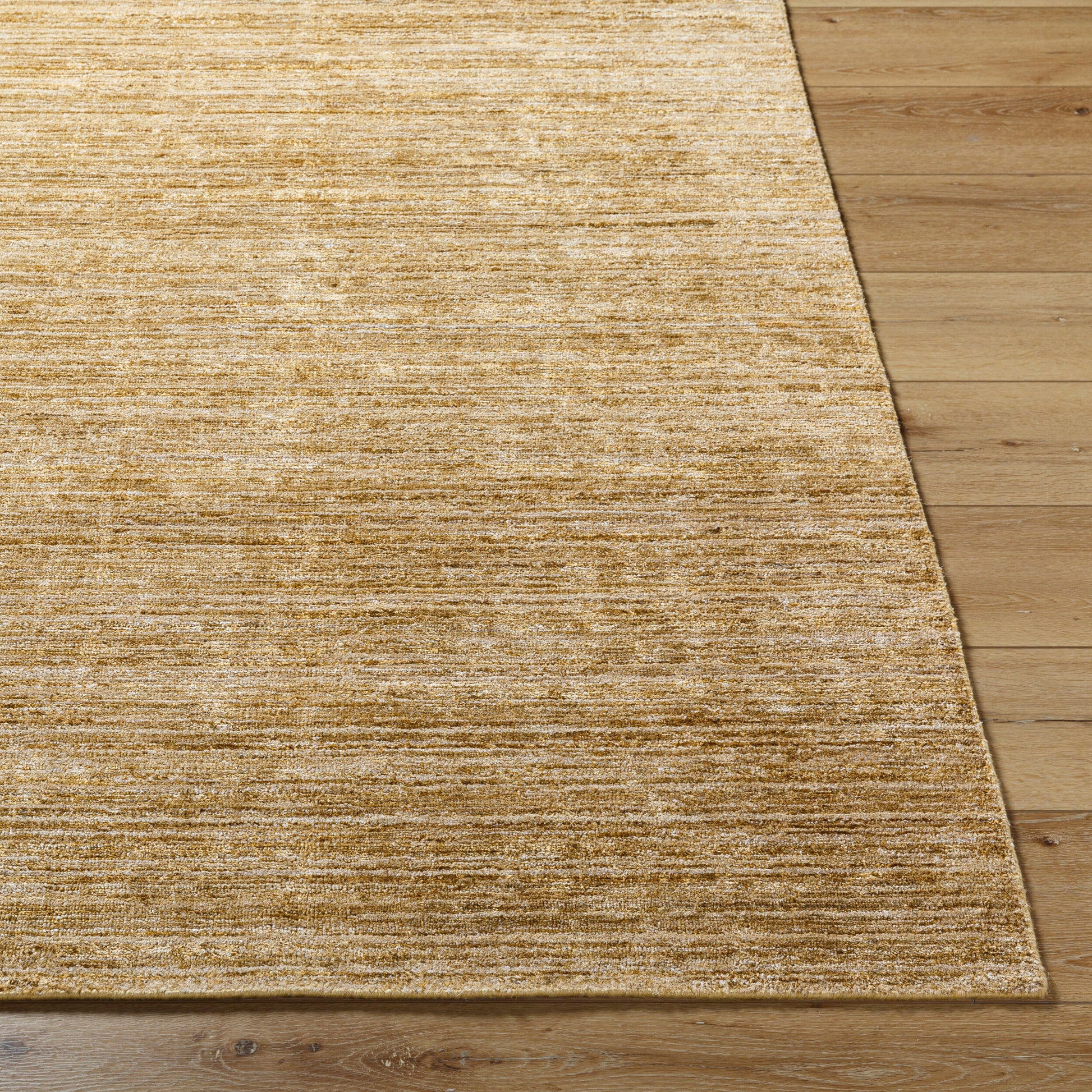 A close-up of the Striated Viscose - 8 x 10 rug, hand knotted in light brown, placed on a wooden floor, highlights its textured surface at the corner next to light wood planks.