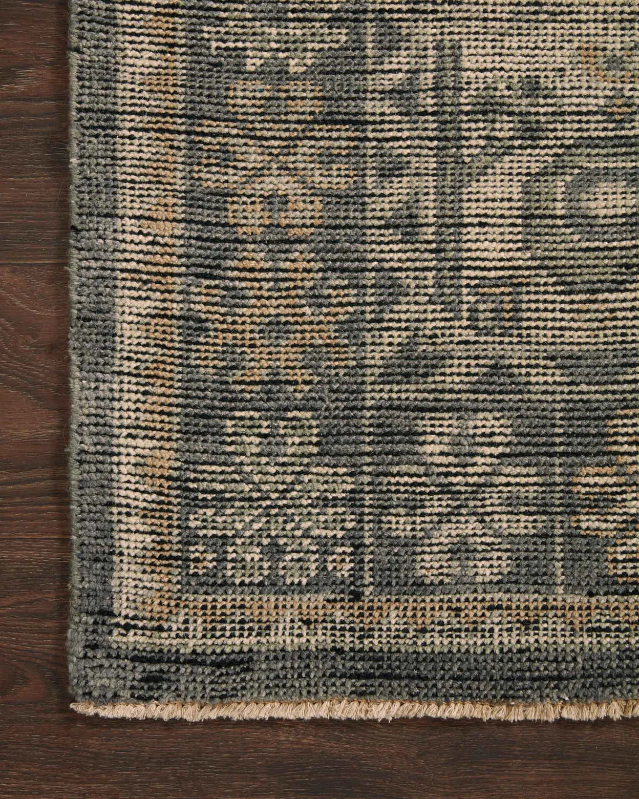 Close-up of the Wool Bone-Charcoal Rug 8'x10', featuring a dark, intricate floral and geometric pattern with a thick textured border, shown on a dark wooden floor. This woven rug is available in various sizes.