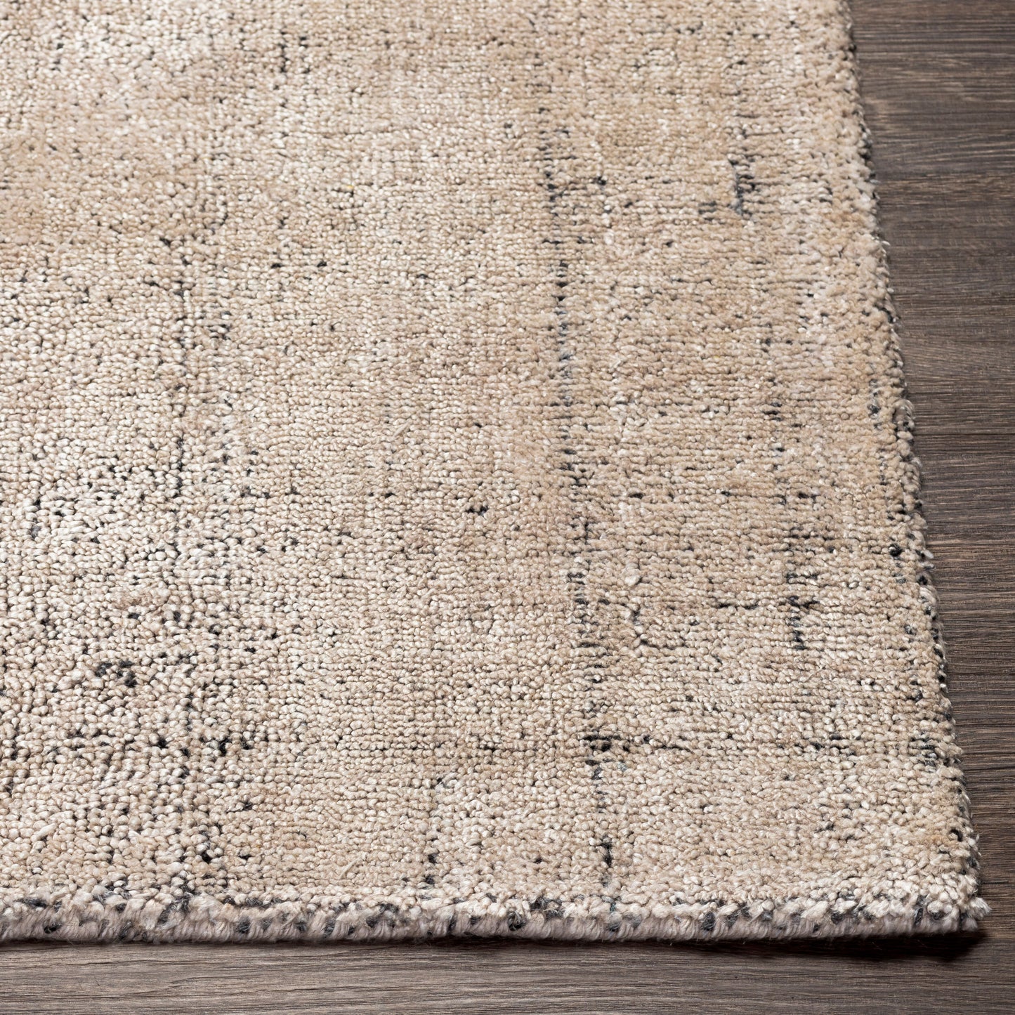 Close-up of the corner of the Silky Hue Wool Rug - 8 x 10 in beige and light brown with subtle dark speckles, featuring a slightly uneven, handmade look, shown on a dark wood floor.