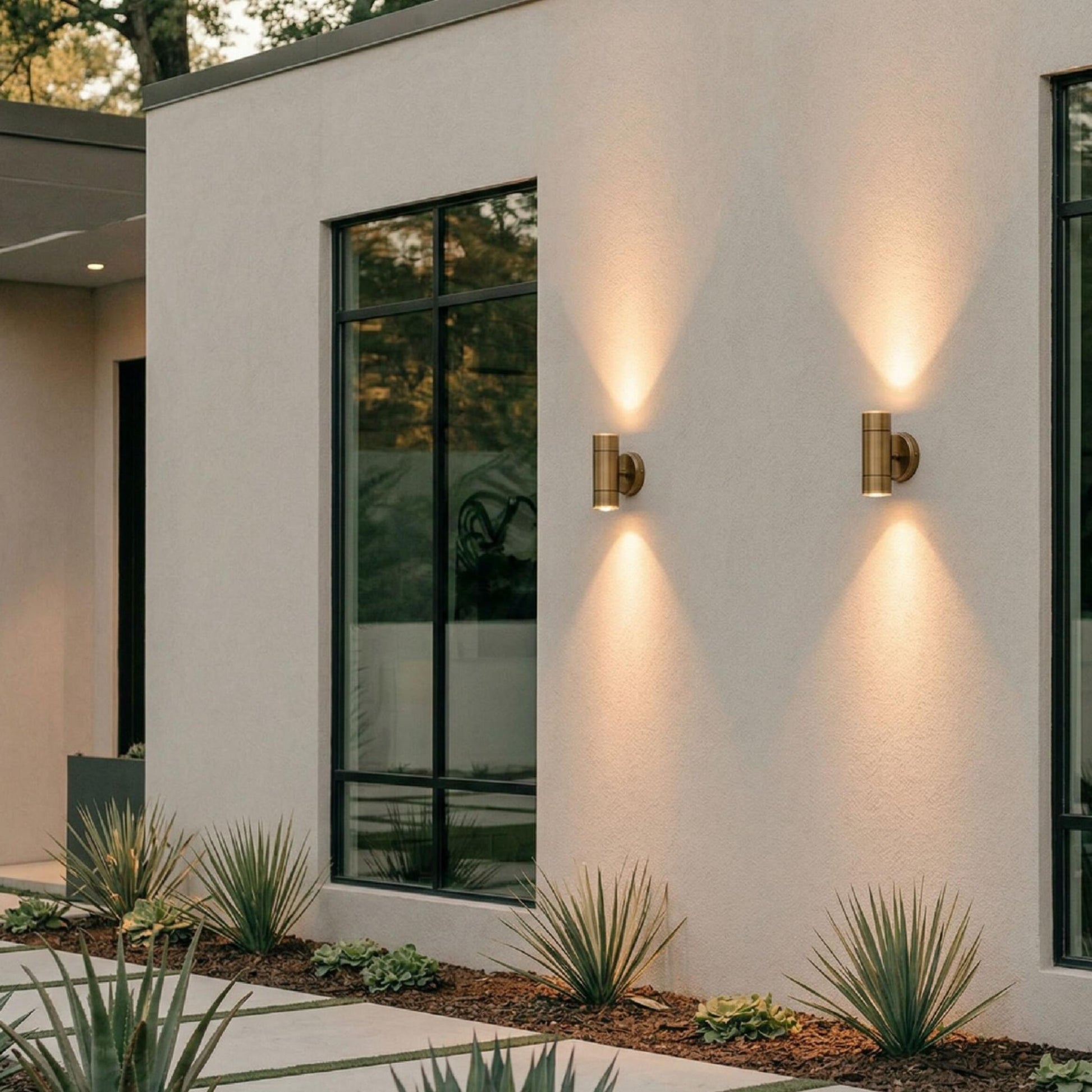A modern exterior wall features two Outdoor Brass Sconces casting light both upward and downward, accompanied by tall black-framed windows, minimalist landscaping with agave plants, a concrete pathway, and a decorative vase thoughtfully positioned by the entryway.
