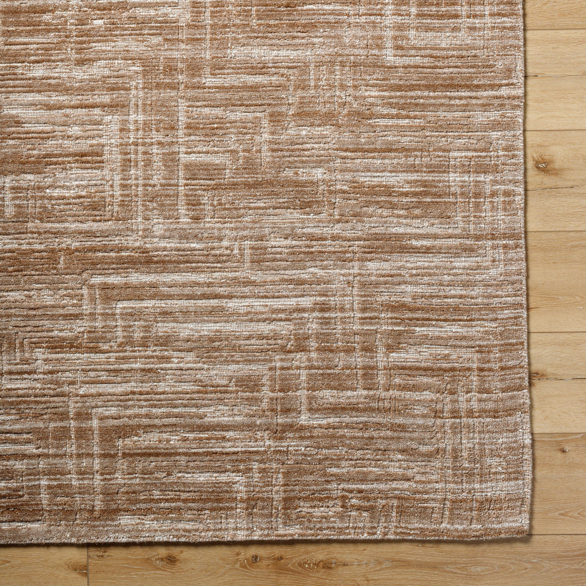 A Handmade Geo Pattern Rug (8'x10') featuring a beige and white abstract linear design is shown on a light wooden floor, with the image capturing the rug's corner and part of the flooring.