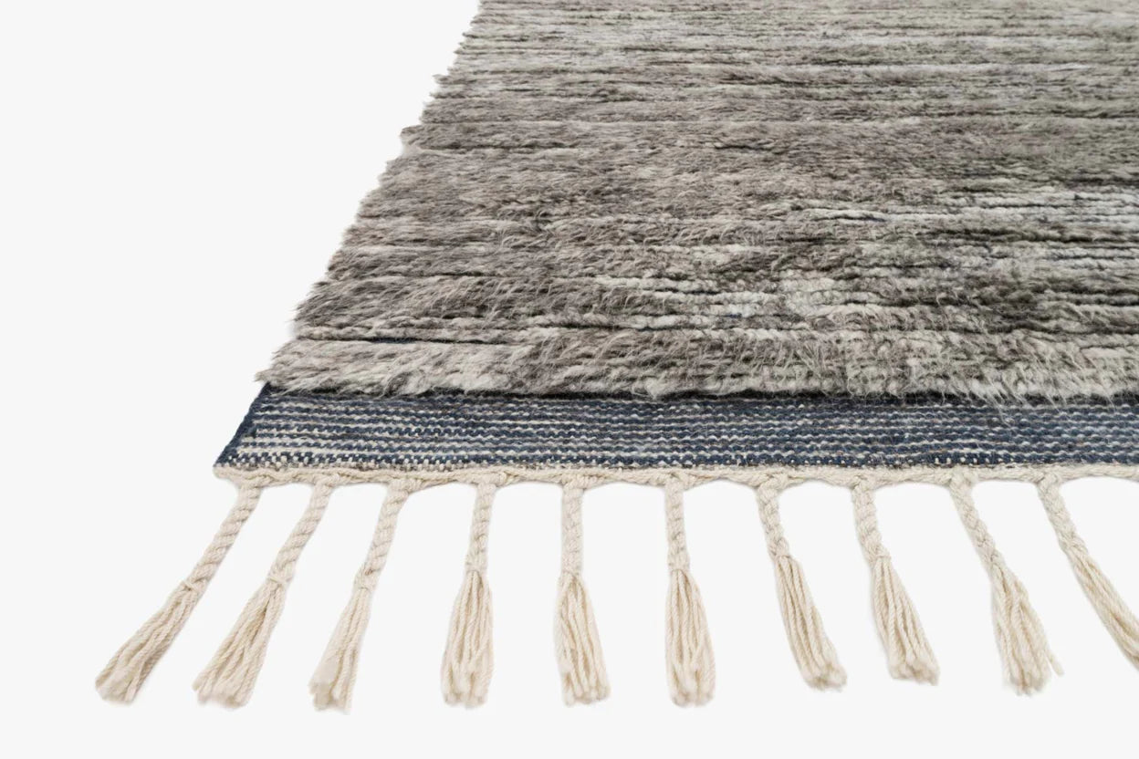 A close-up of the Indian Wool Rug With Fringes - 8'6x11'6, featuring a gray and black textured shaggy surface, blue woven edge, and cream fringe tassels on one side. Presented against a white background.