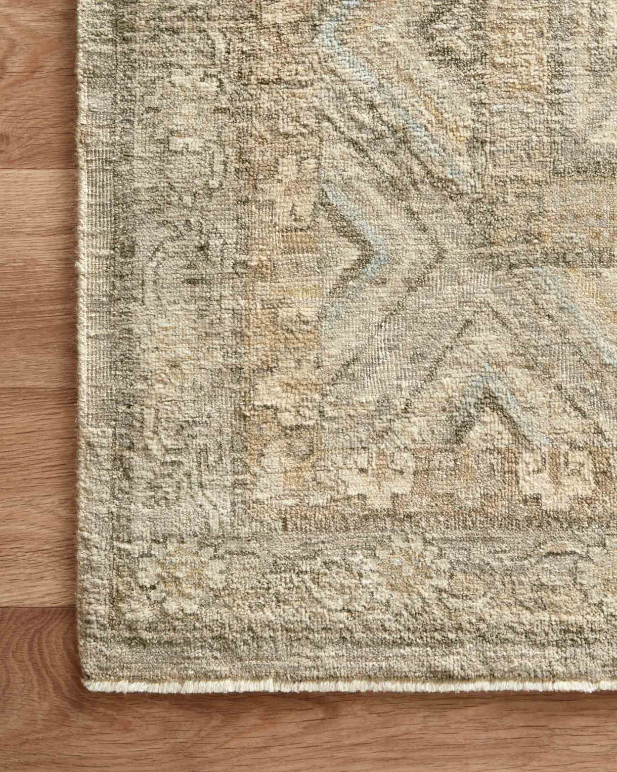 A close-up of the corner of the Hand Knotted Ash Rug 8'x10', featuring a textured, faded geometric pattern in beige and taupe, shown on a light wooden floor.