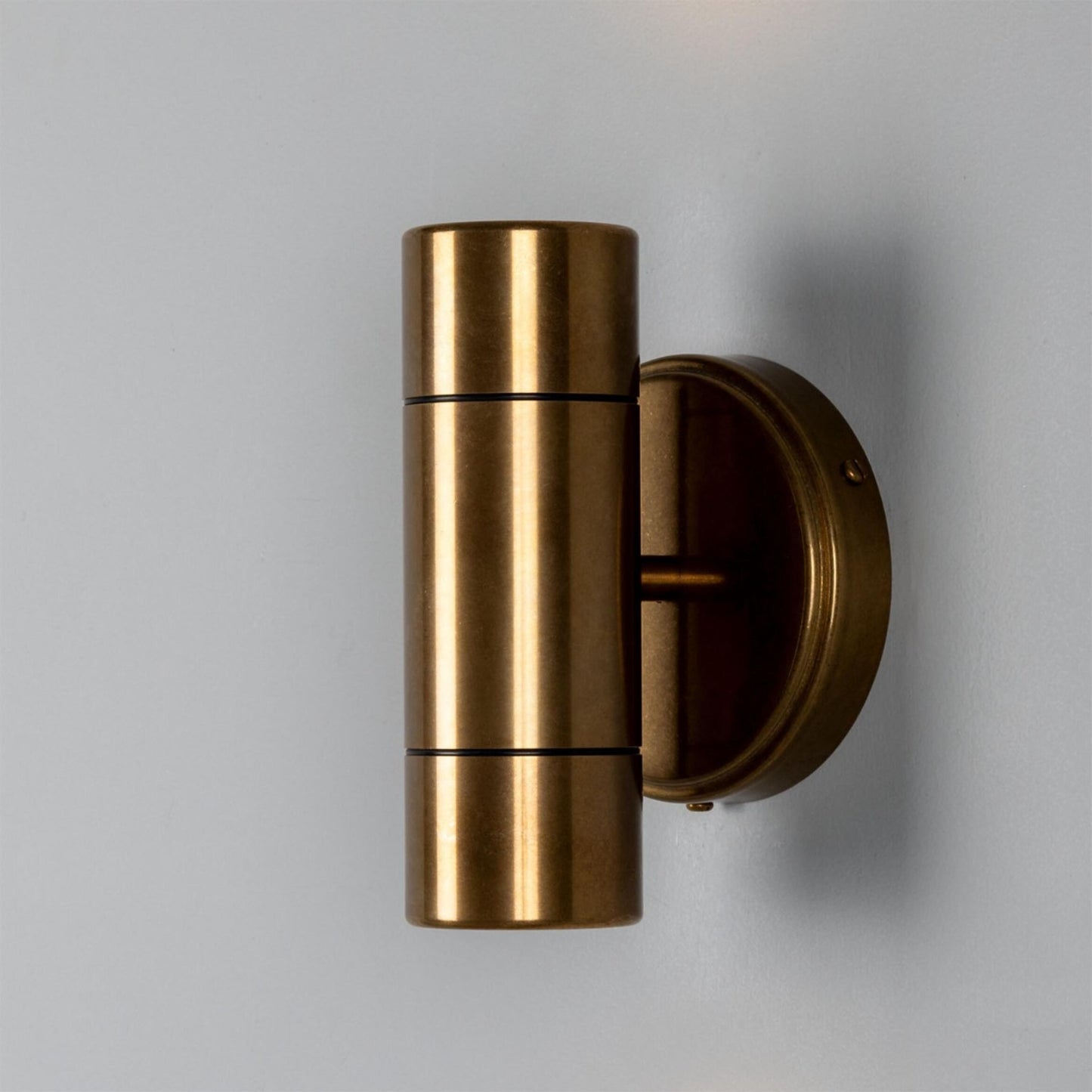 The Outdoor Brass Sconce—a cylindrical brass wall fixture with top and bottom light sources, mounted on a round backplate against a simple gray wall—is paired elegantly with a decorative vase to enhance any modern interior.