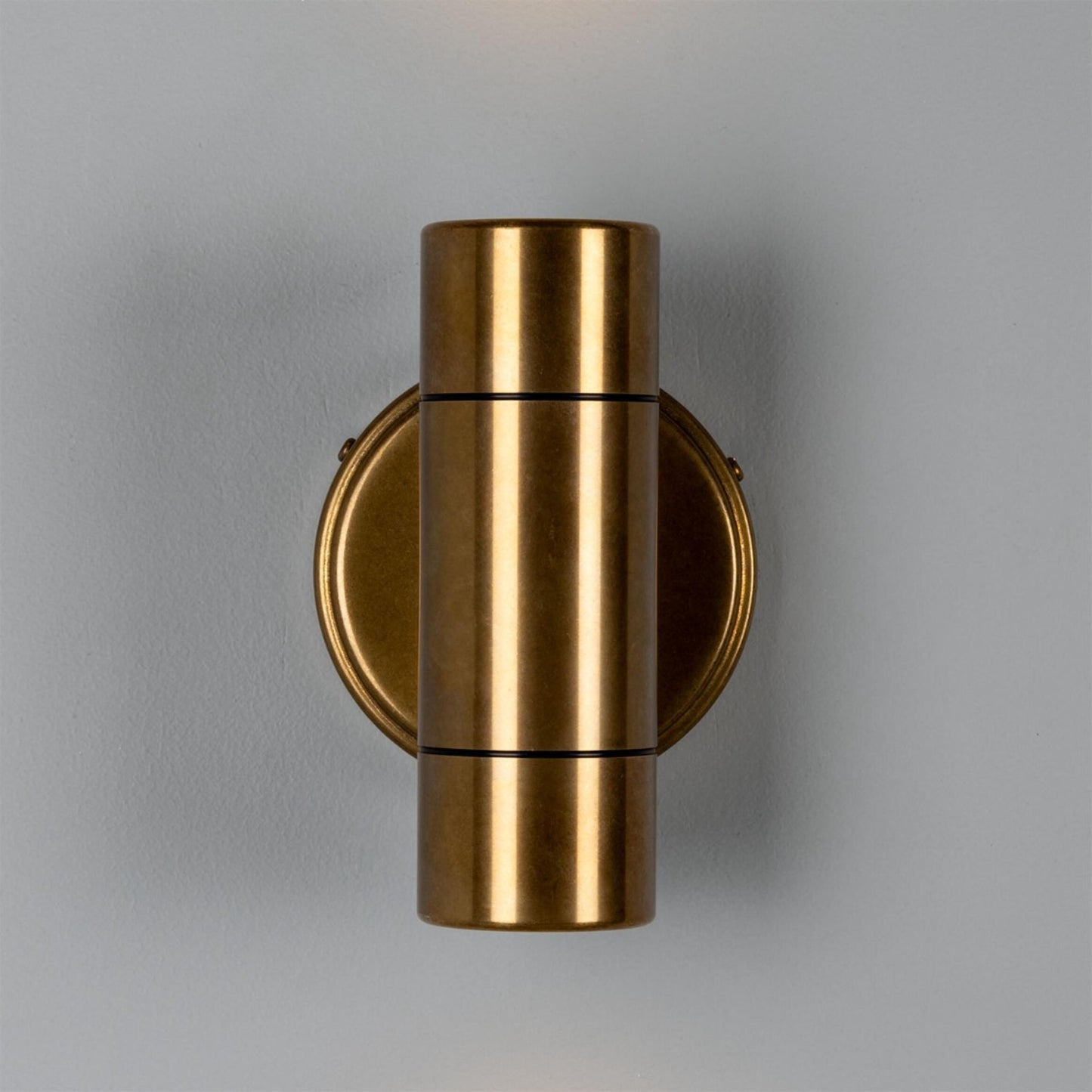 The Outdoor Brass Sconce, featuring a cylindrical brass design mounted on a round brass base, casts a soft glow from its top and bottom edges against a light gray wall. Pair this elegant fixture with a decorative vase nearby to create a refined and cohesive look in any space.