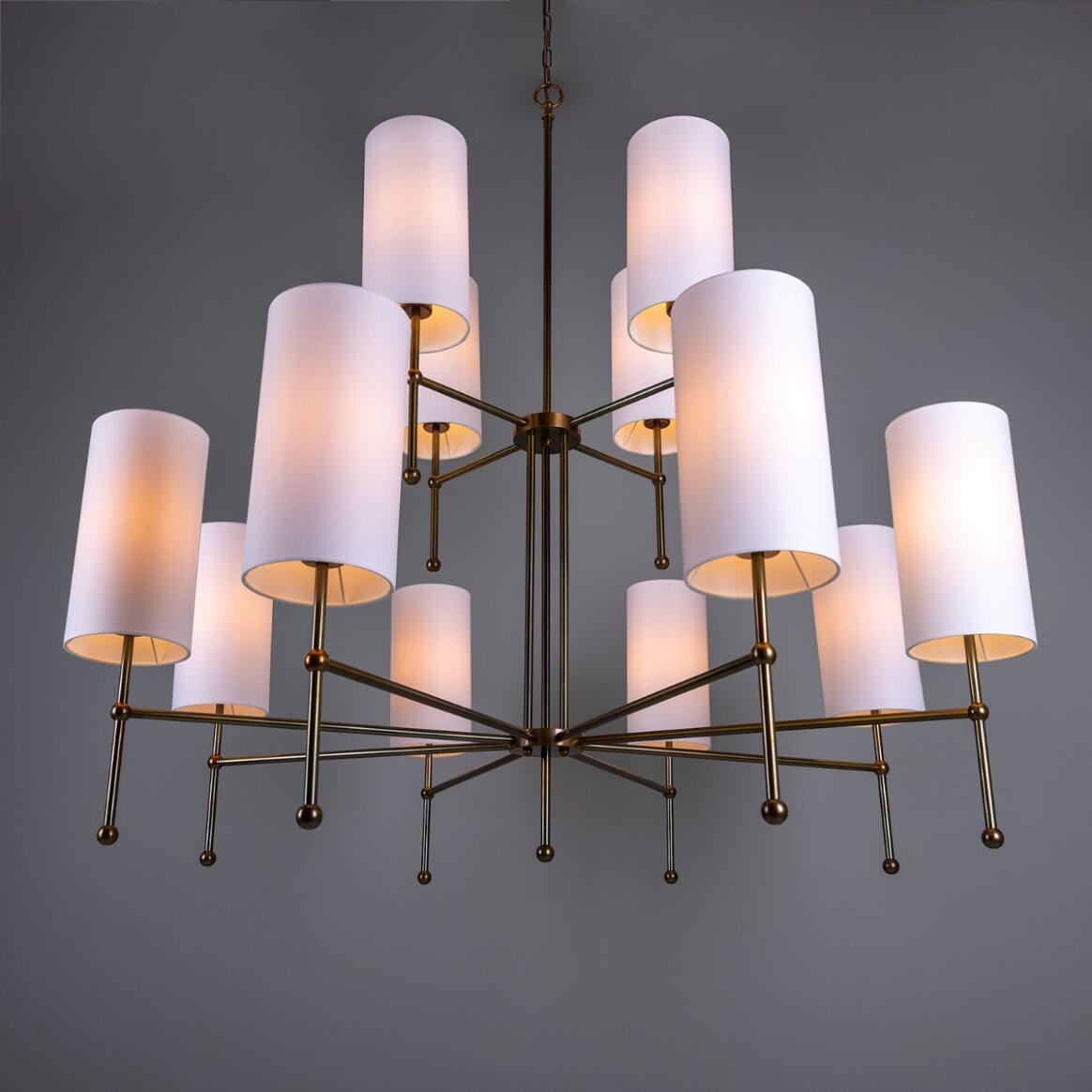 Introducing the Sonoran Chandelier—a modern, contemporary fixture featuring a bronze frame and ten cylindrical white lampshades. This sophisticated multi-tier chandelier brings timeless style to any space, hanging elegantly against a plain gray backdrop and perfectly blending classic design with modern appeal.