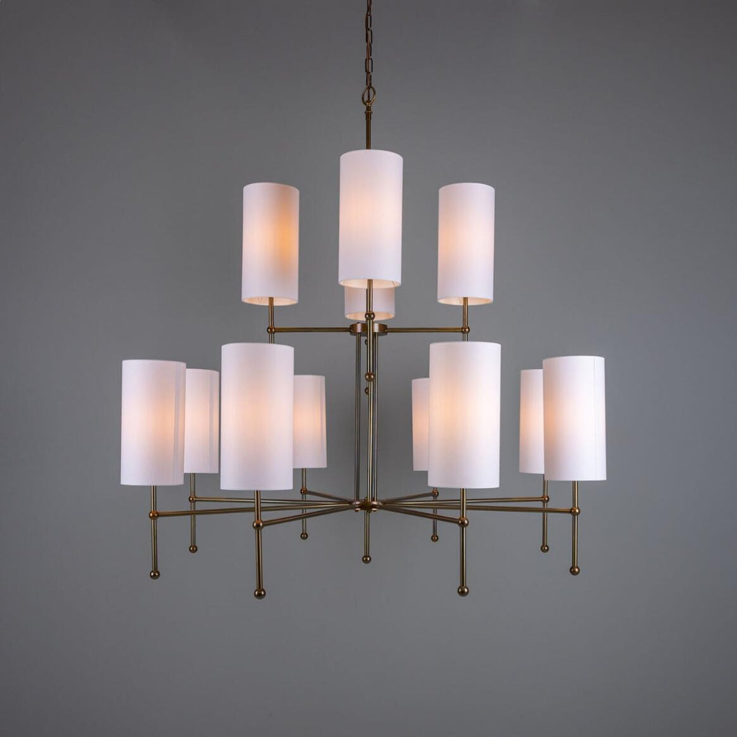 The Sonoran Chandelier features twelve cylindrical white lampshades arranged in two tiers and suspended from the ceiling by a chain, set against a plain gray background—a striking example of multi-tier lighting for contemporary interiors.