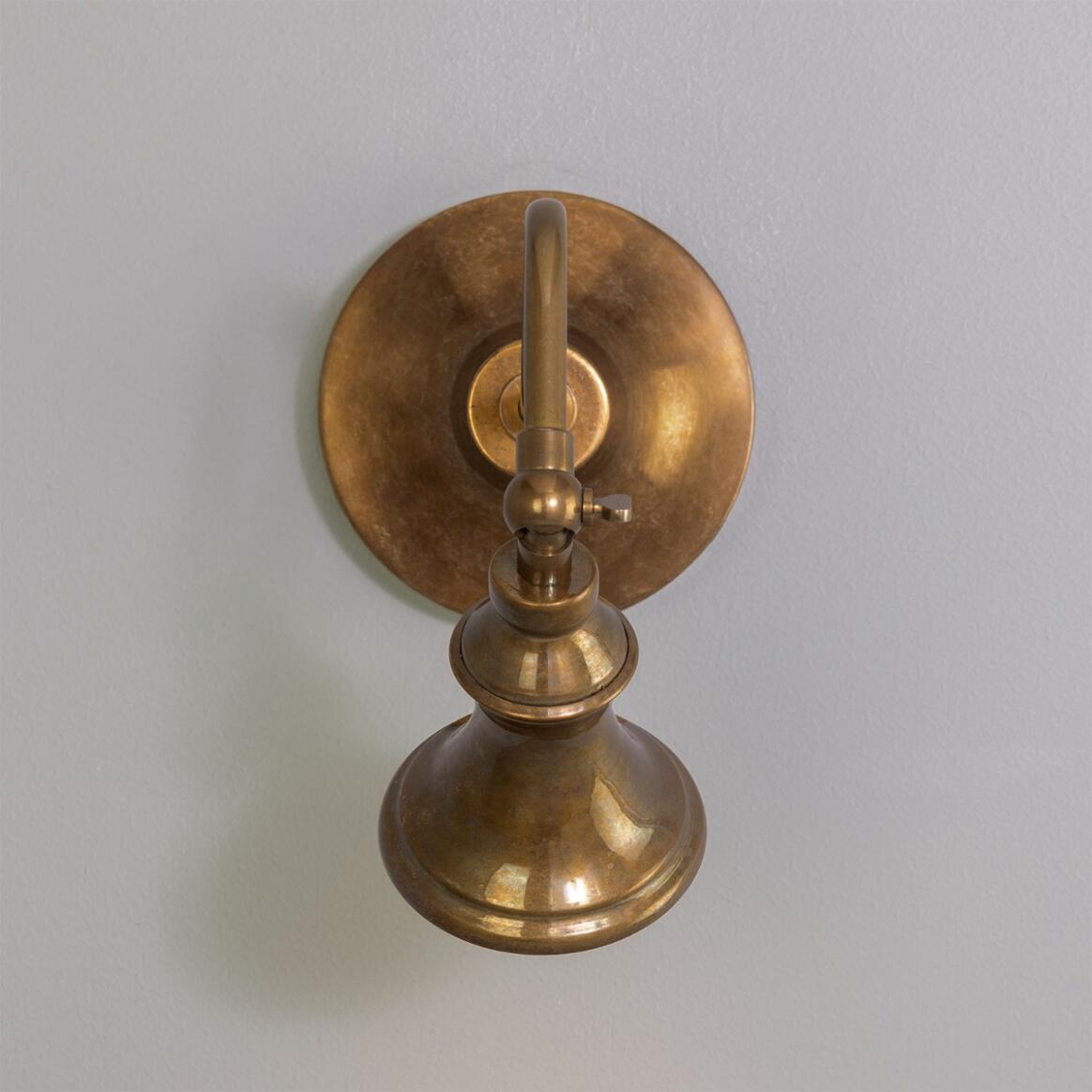 The Adjustable Bell Sconce, showcasing a classic design with a round base and curved arm in antique brass, is beautifully mounted on a light gray wall.